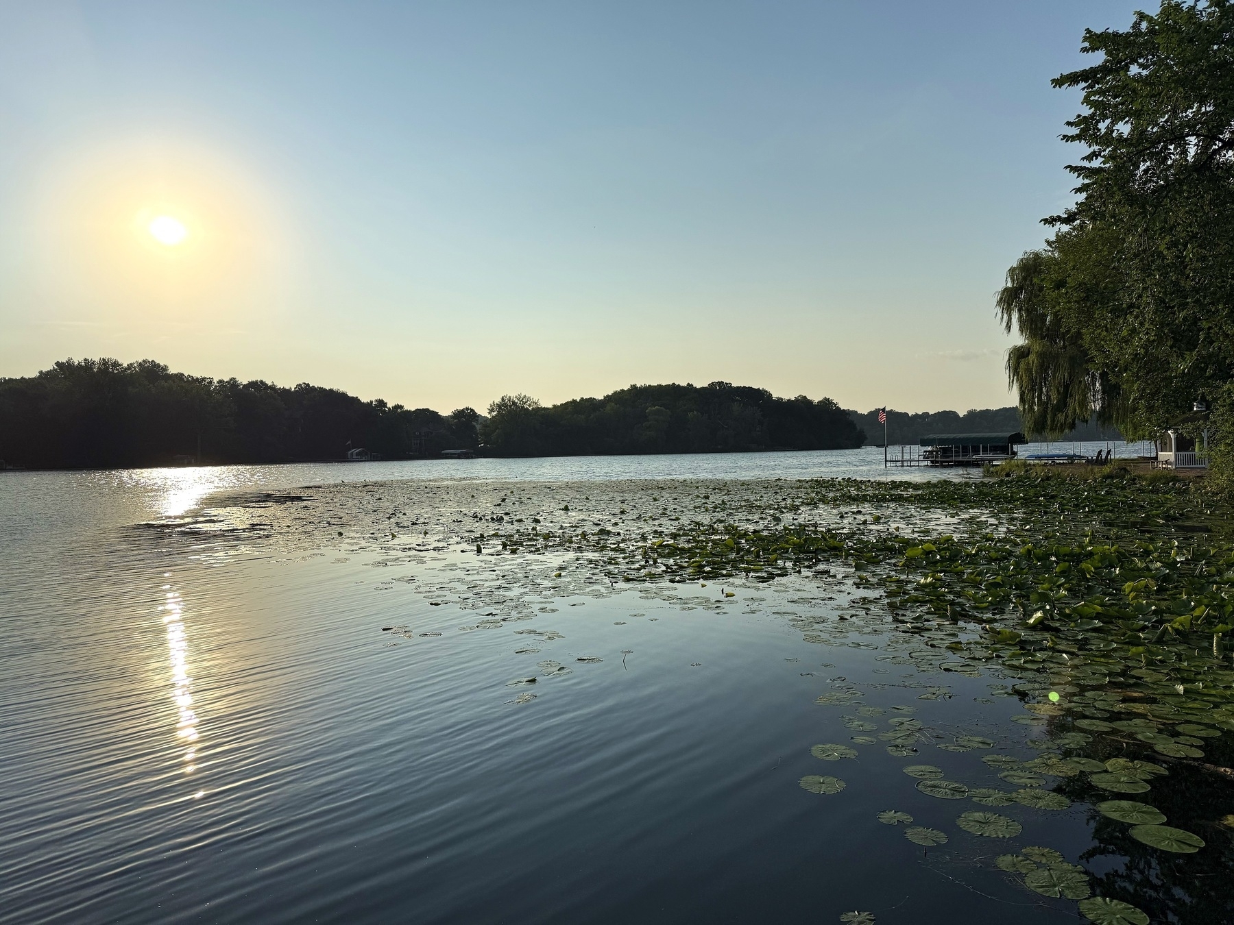 A serene lake at sunset with sunlight reflecting on the water. The surface is dotted with lily pads, and trees line the shore. A small pier with a flag is visible in the background.