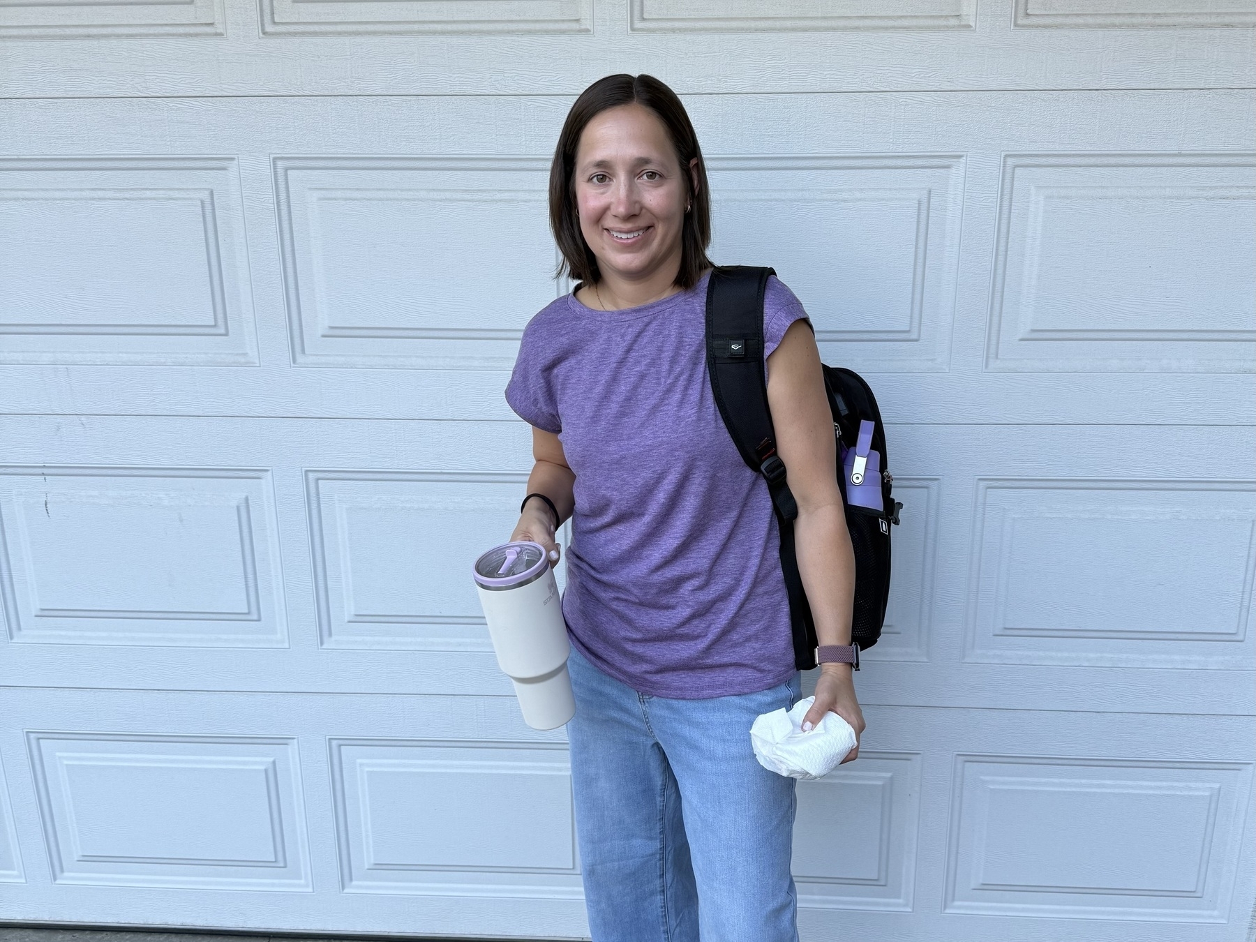 A person stands in front of a garage door, wearing a purple shirt and light blue jeans. They carry a black backpack with a pen attached and hold a white travel mug and a paper towel.