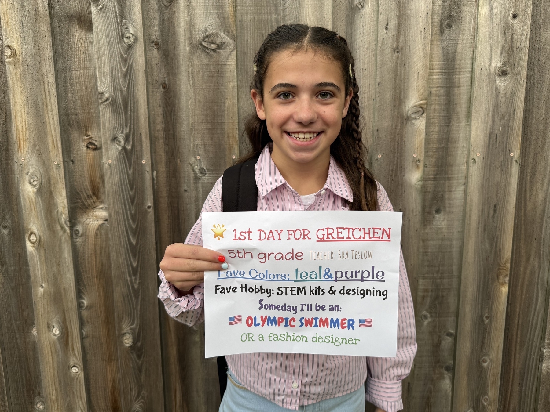 Girl holding a sign that reads: “1st day for Gretchen, 5th grade. Teacher: Sra Teslow. Fave colors: teal & purple. Fave hobby: STEM kits & designing. Someday I’ll be an Olympic swimmer or a fashion designer.” She is smiling,
