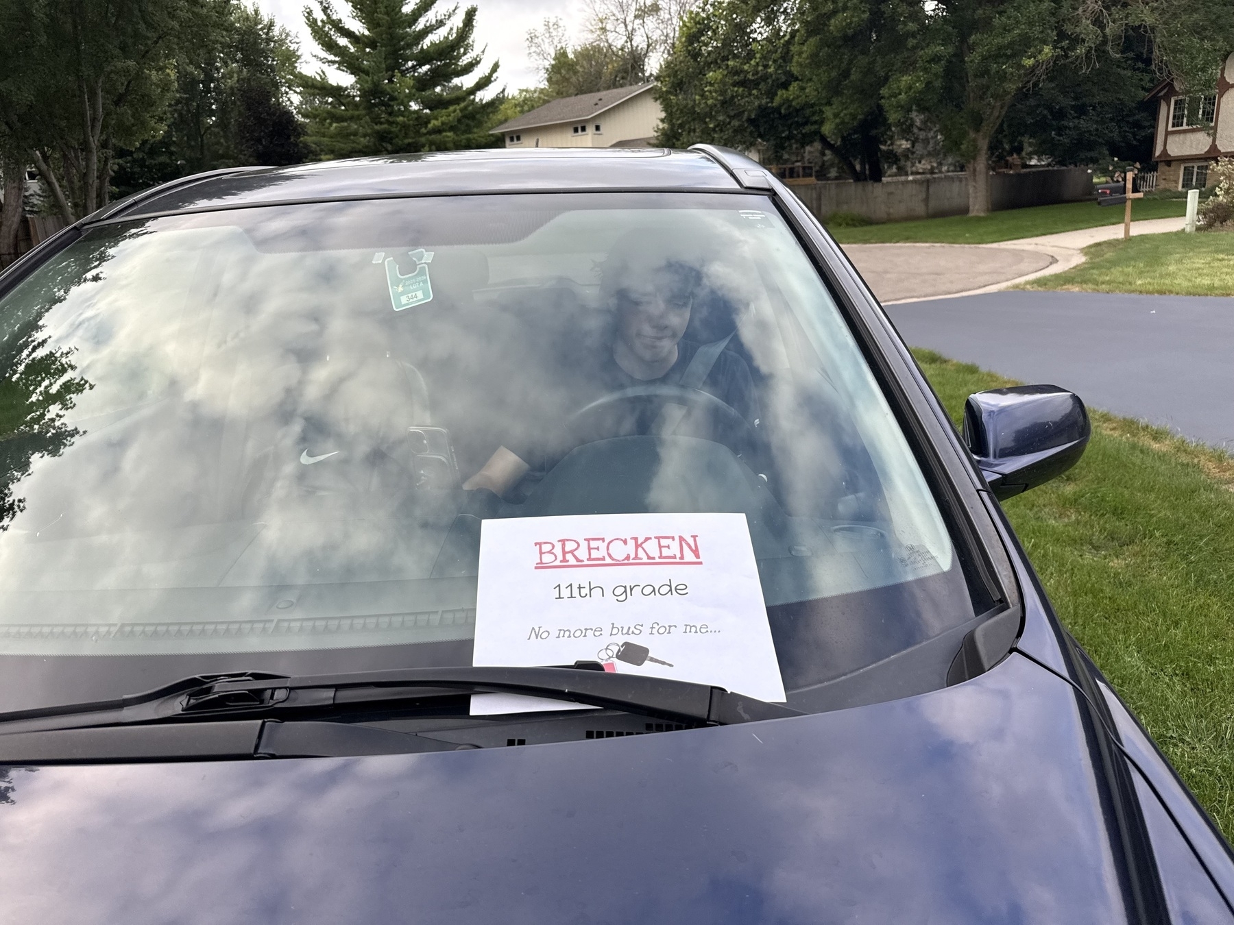 A person sits in the driver’s seat of a car, with a sign on the windshield reading “Brecken 11th grade No more bus for me,” indicating a transition to driving themselves to school. The vehicle is parked on a residential street with trees and grass nearby.