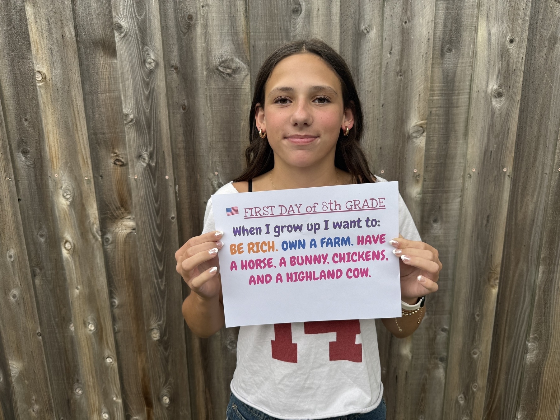 A young girl stands in front of a wooden fence, holding a sign that reads, “First day of 8th grade. When I grow up I want to: be rich, own a farm, have a horse, a bunny, chickens, and a Highland cow.