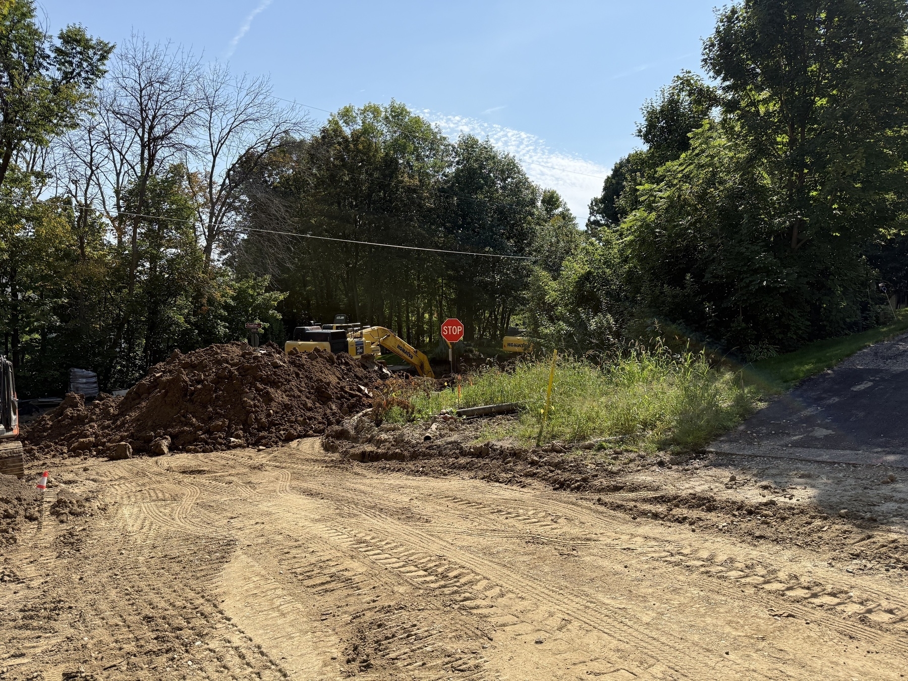 A construction site with dirt mounds and an excavator partially visible behind dense trees. A stop sign is located near the excavator.