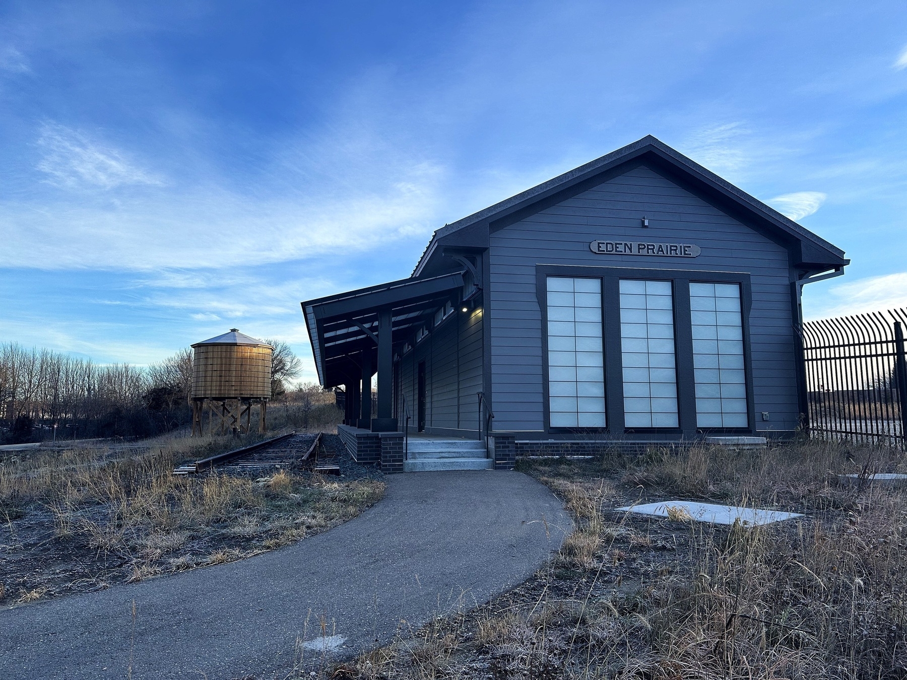 A rustic train depot labeled “Eden Prairie” with a wooden water tower in the background. The building has a wooden exterior with large windows, and a pathway leads up to it. Surrounding the area are grassy fields and a clear blue sky.