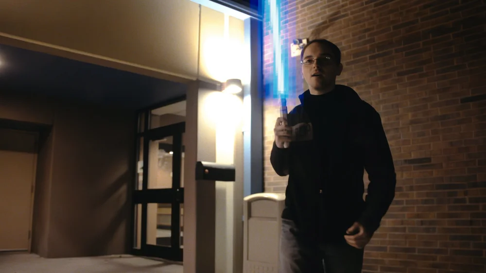 A person in dark clothing is standing outside near a building, holding a glowing blue sword-like object.