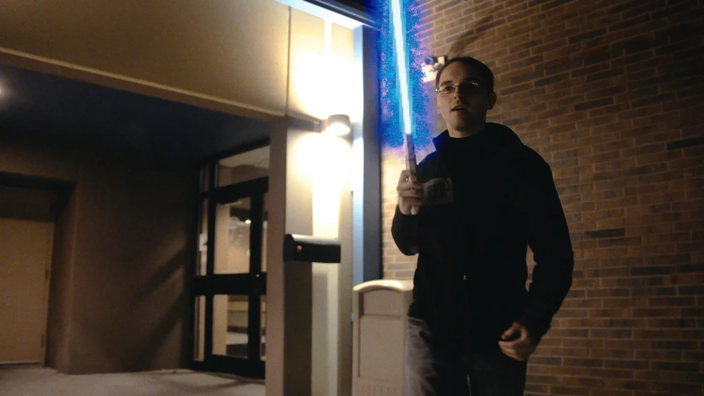 A person is standing outside a building at night, holding a glowing blue lightsaber.