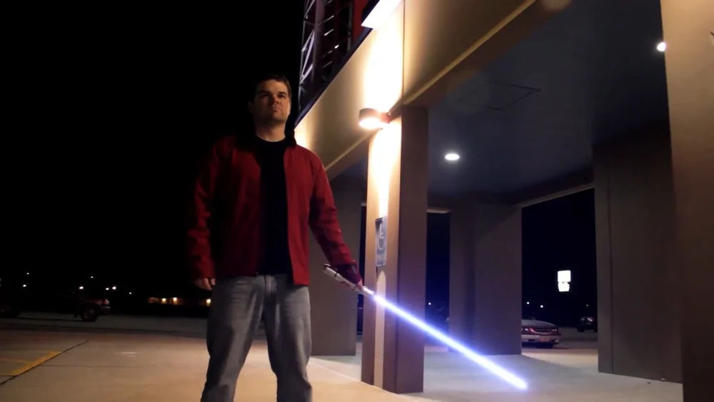 A person stands outside at night holding a glowing lightsaber-like object under a building with illuminated columns.