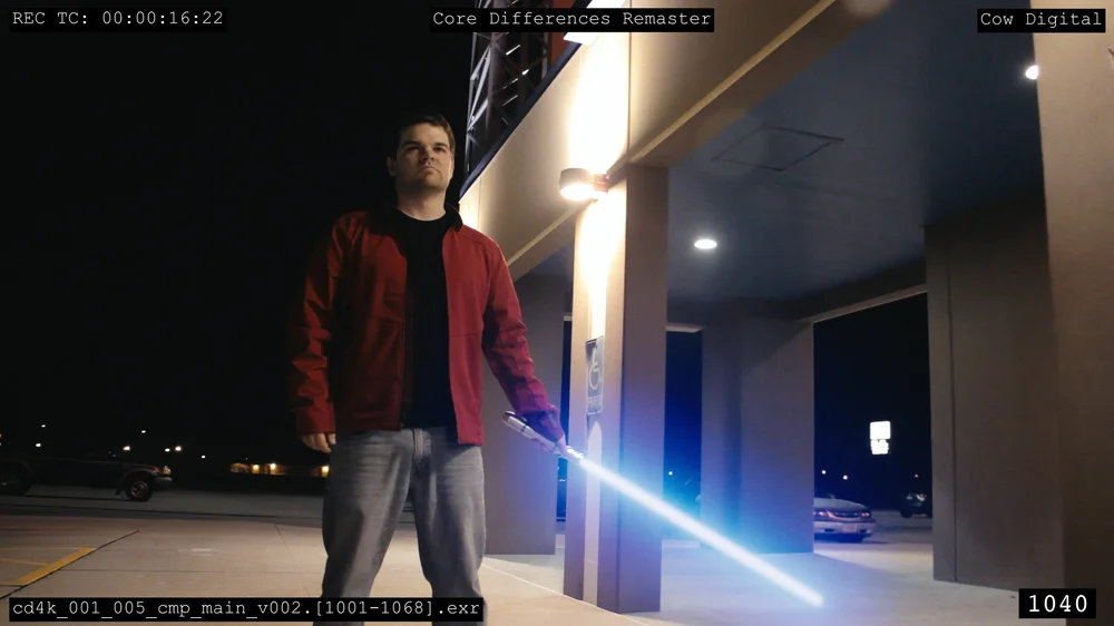 A person wearing a red jacket holds a glowing blue lightsaber while standing outside near a building at night.