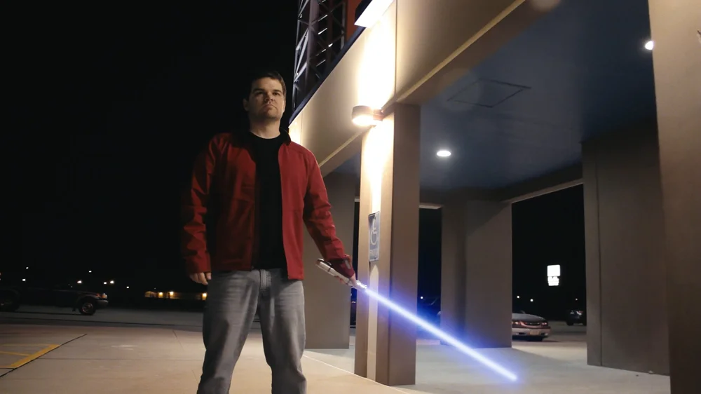 A person standing outside under a lit awning is holding a glowing blue lightsaber.