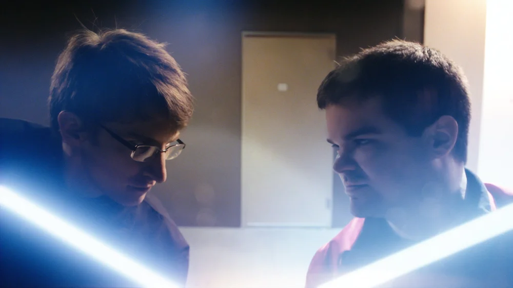Two individuals are facing each other with glowing lightsabers.