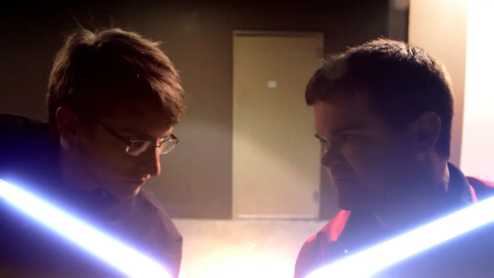 Two people are facing each other with glowing lightsaber-like objects in a dimly lit room.
