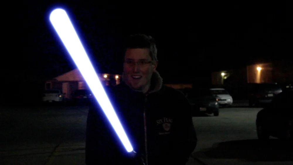 A person holds a brightly lit, white lightsaber at night in a dimly lit environment.