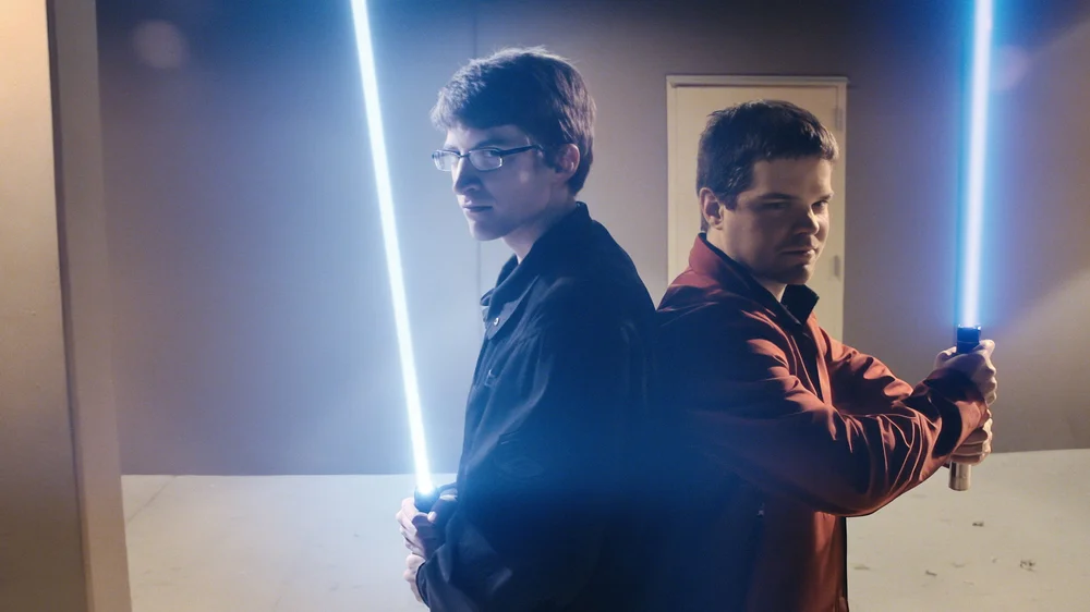 Two individuals stand back-to-back holding illuminated lightsabers in a dimly lit room.