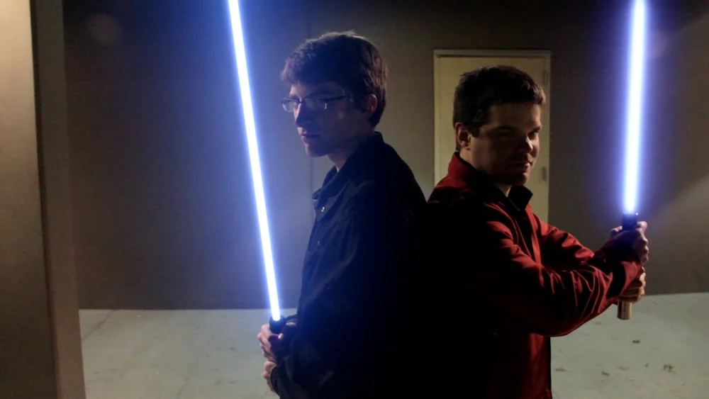 Two individuals are standing back to back, each holding a lit lightsaber, one blue and the other red.