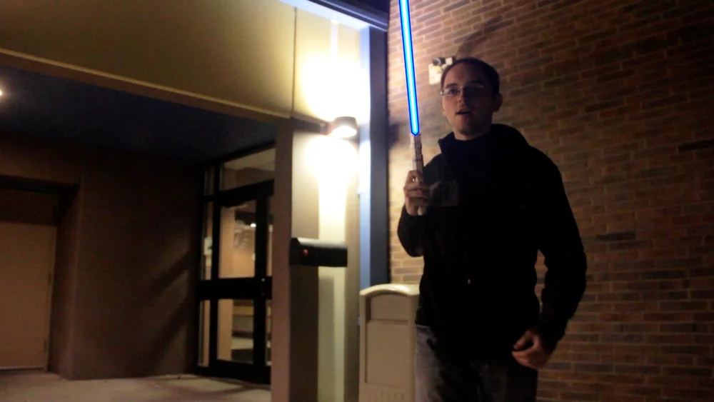 A person holding a glowing blue lightsaber stands outside near a building entrance at night.
