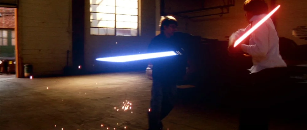 Two people are engaged in a duel with glowing, saber-like weapons inside a dimly lit room.