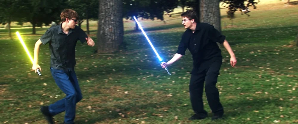 Two individuals are playfully engaging in a lightsaber duel in a grassy park setting.