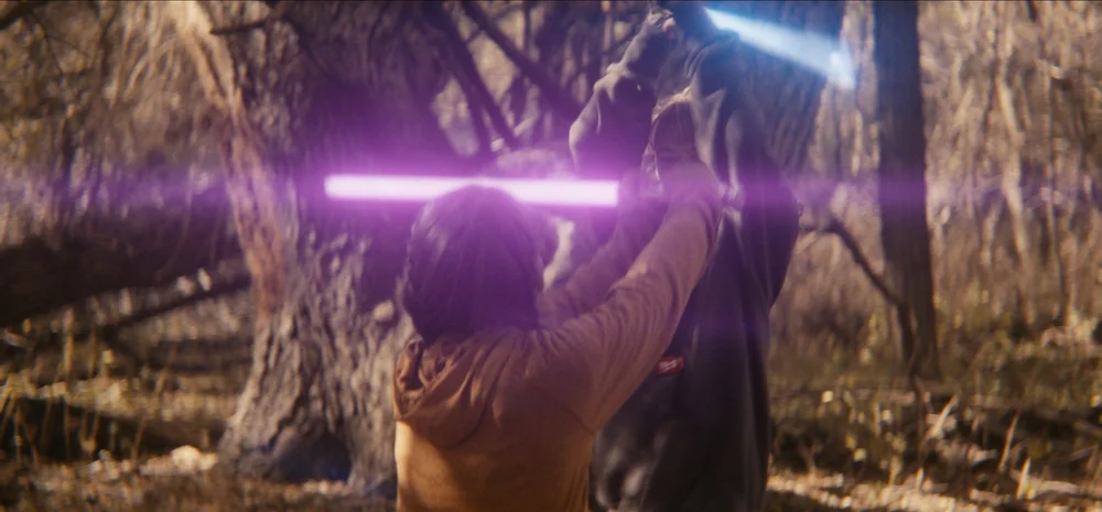 Two individuals are engaged in a lightsaber duel in a forest setting.