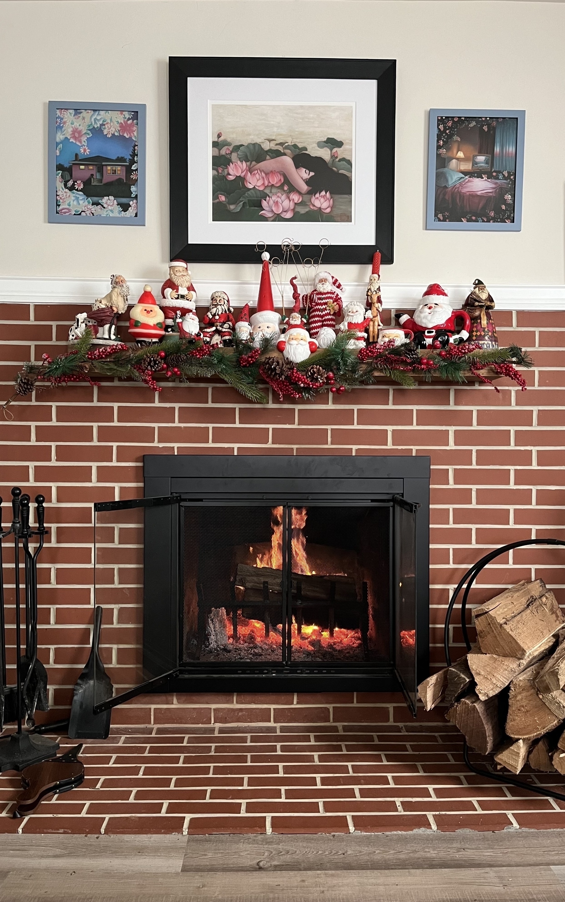 Fireplace with fire burning. A stack of wood to the right of the frame. A mantle full of 15 Santa Clauses with holly and berries. 