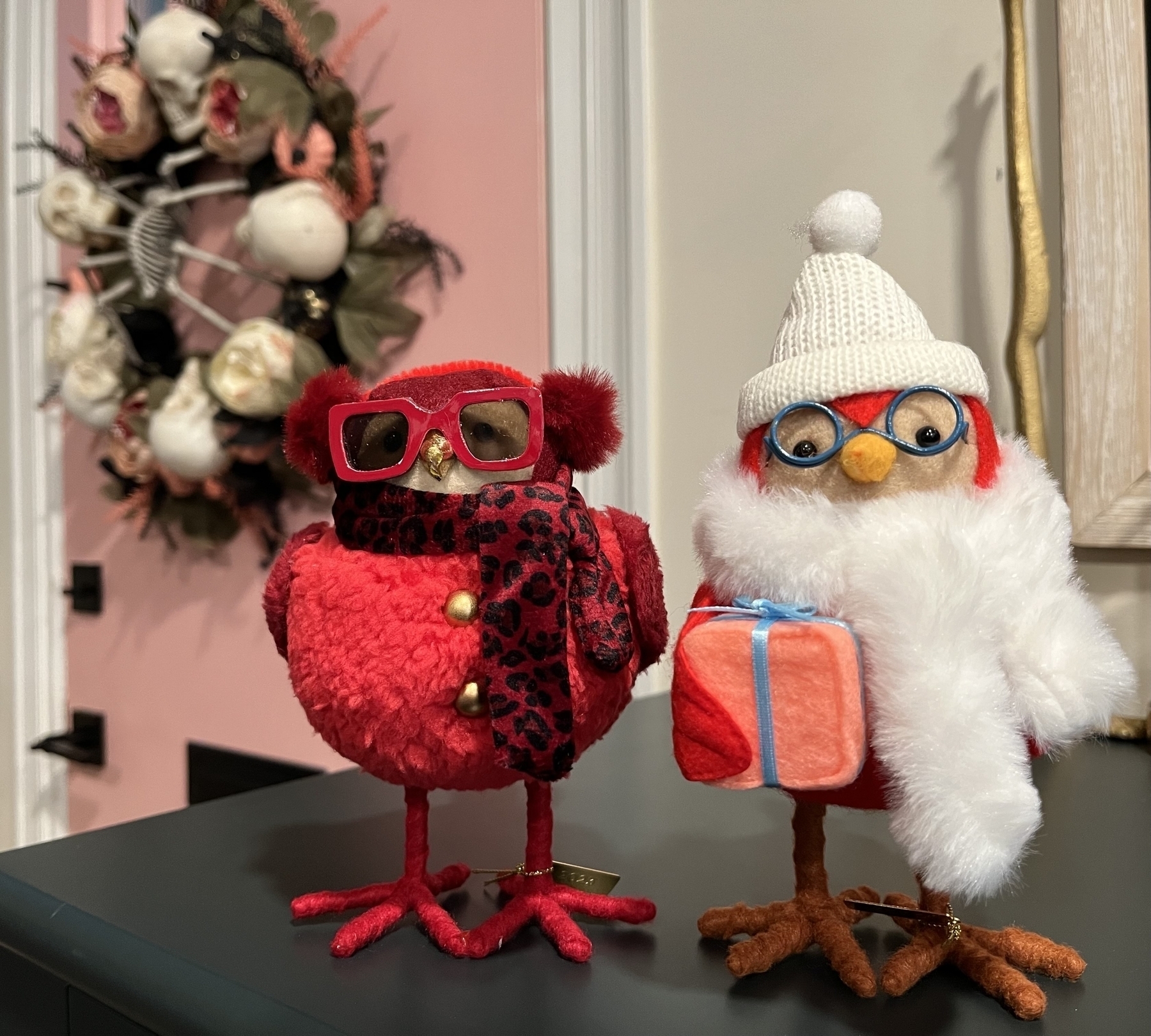 Ruby, a red bird with large red sunglasses and a red scarf standing next to Ginger, a red bird in a white hat and white scarf holding a wrapped present. 