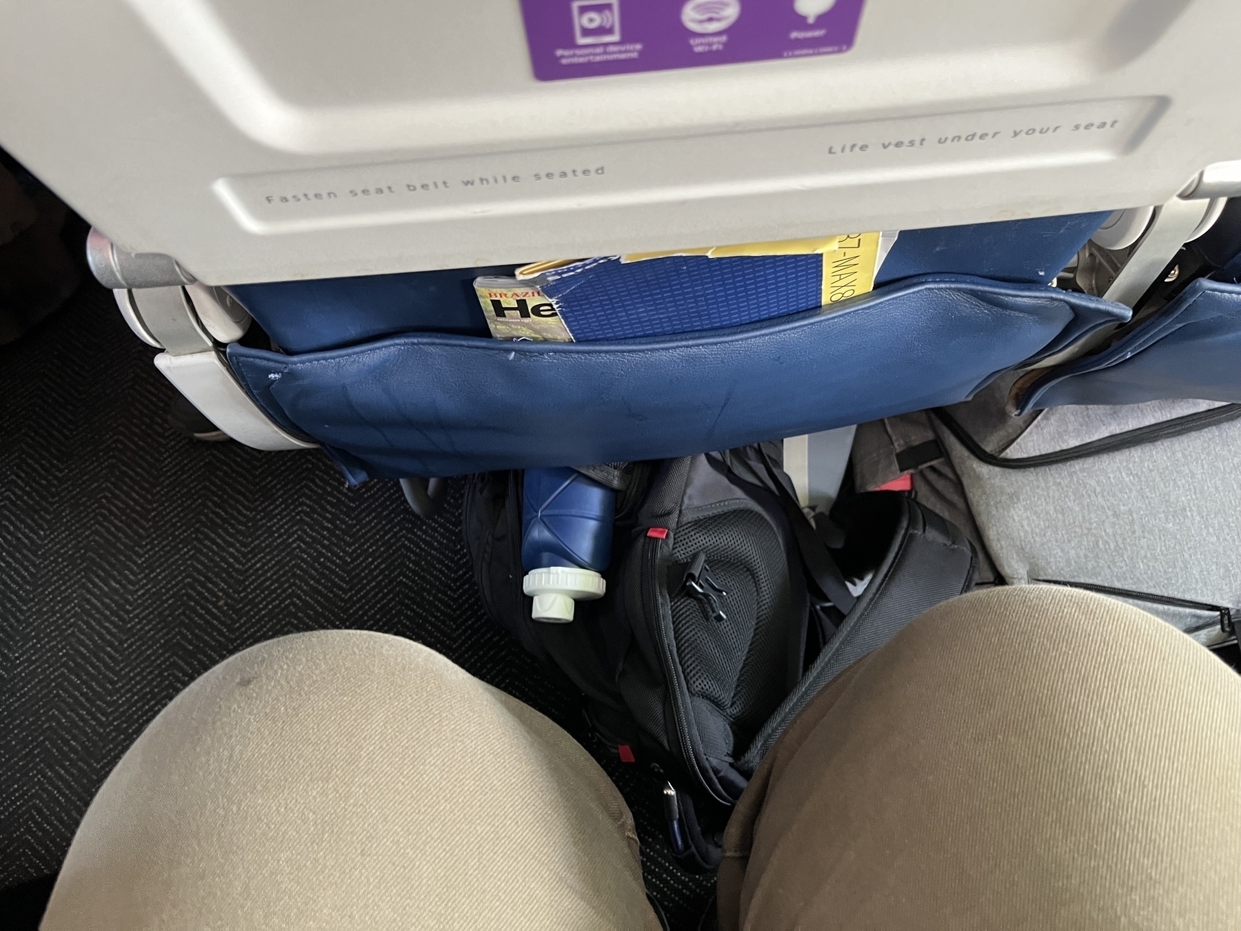 Pair of legs with 6” between knees and seat with a backpack visible on the ground and tray table up. 