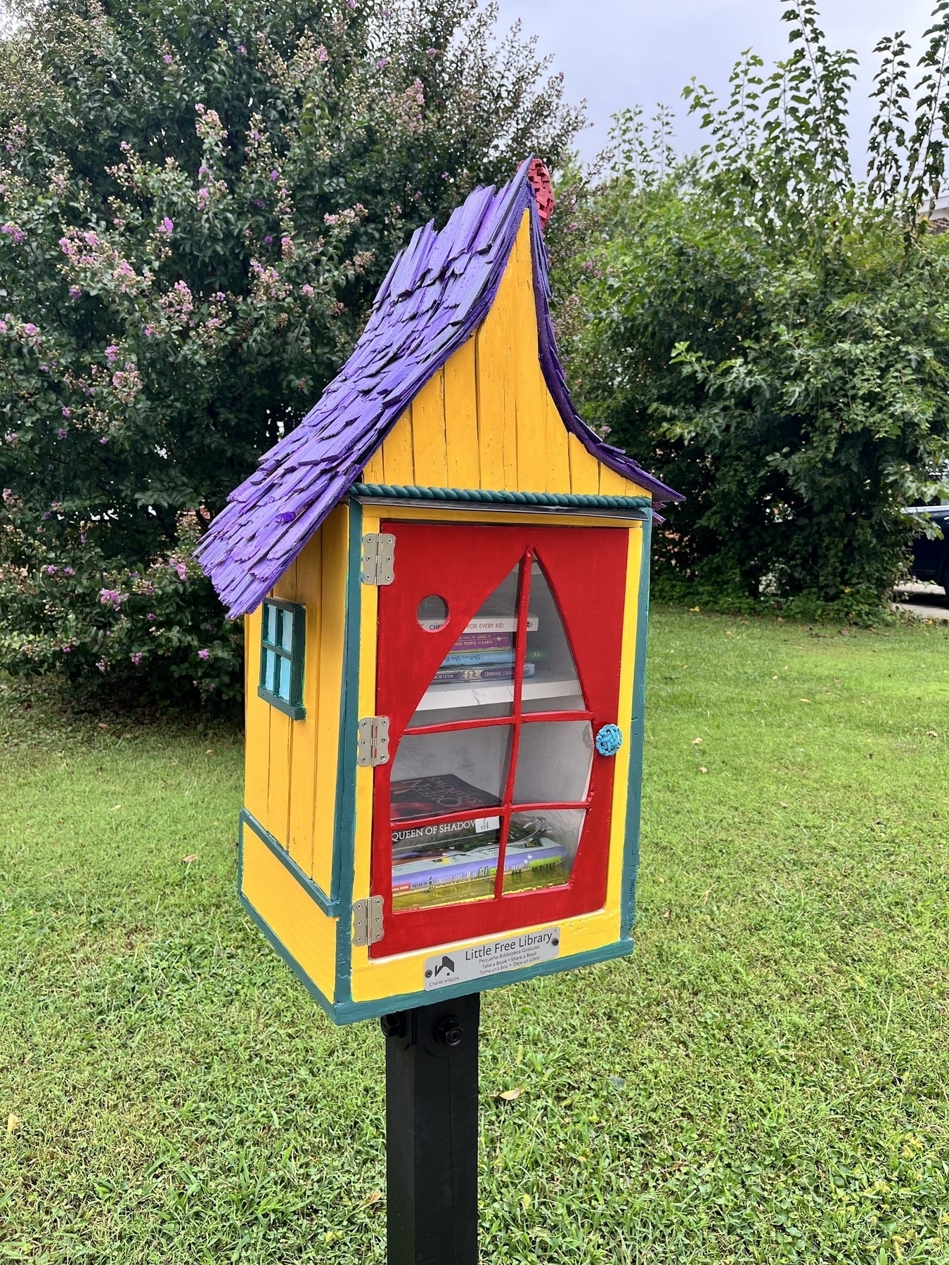 Little Free Library painted yellow with a purple shingle roof.  