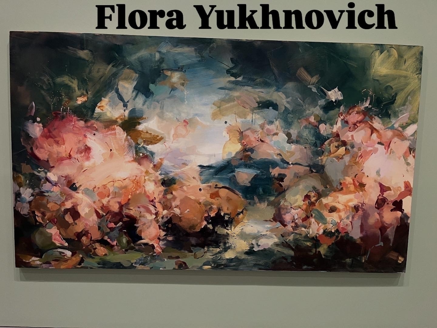 Painting depicts abstract floral forms in pink, orange, and green, blending seamlessly. Located in an art gallery, signed “Flora Yukhnovich” above. The brushstrokes are fluid and dynamic.