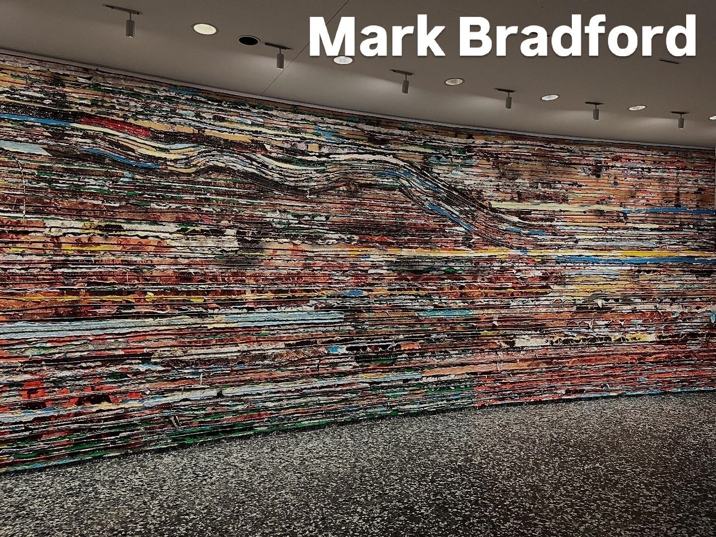 A large, textured abstract mural stretches across a curved wall, featuring layered horizontal strips of colorful paint. The setting includes a patterned floor and overhead spotlights. Text reads: “Mark Bradford.