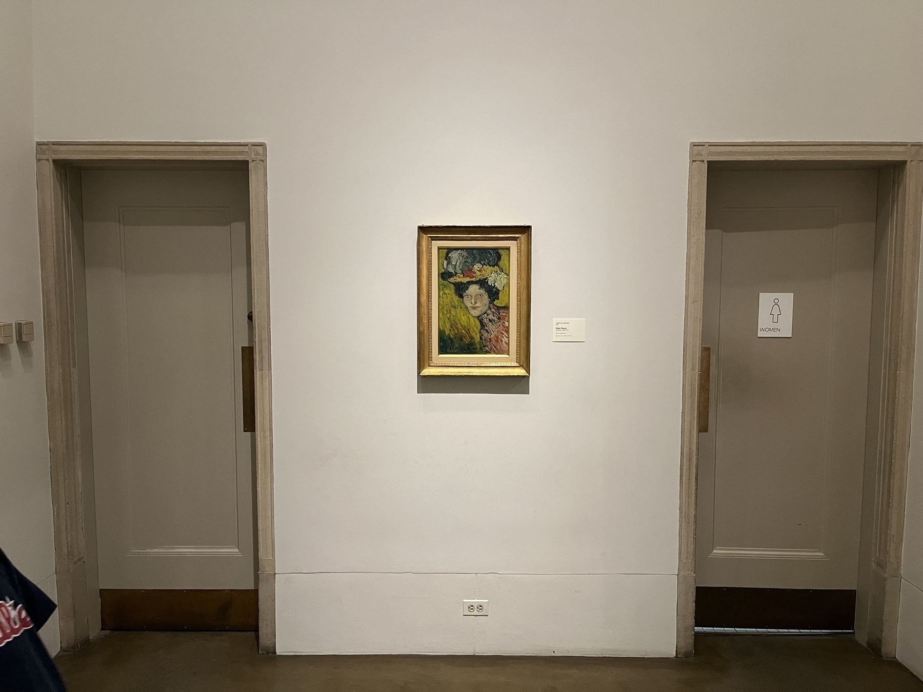 A framed painting by Picasso is displayed on a museum wall between two closed restroom doors. The door on the right is labeled "WOMEN." The painting features a person with a hat and colorful background.