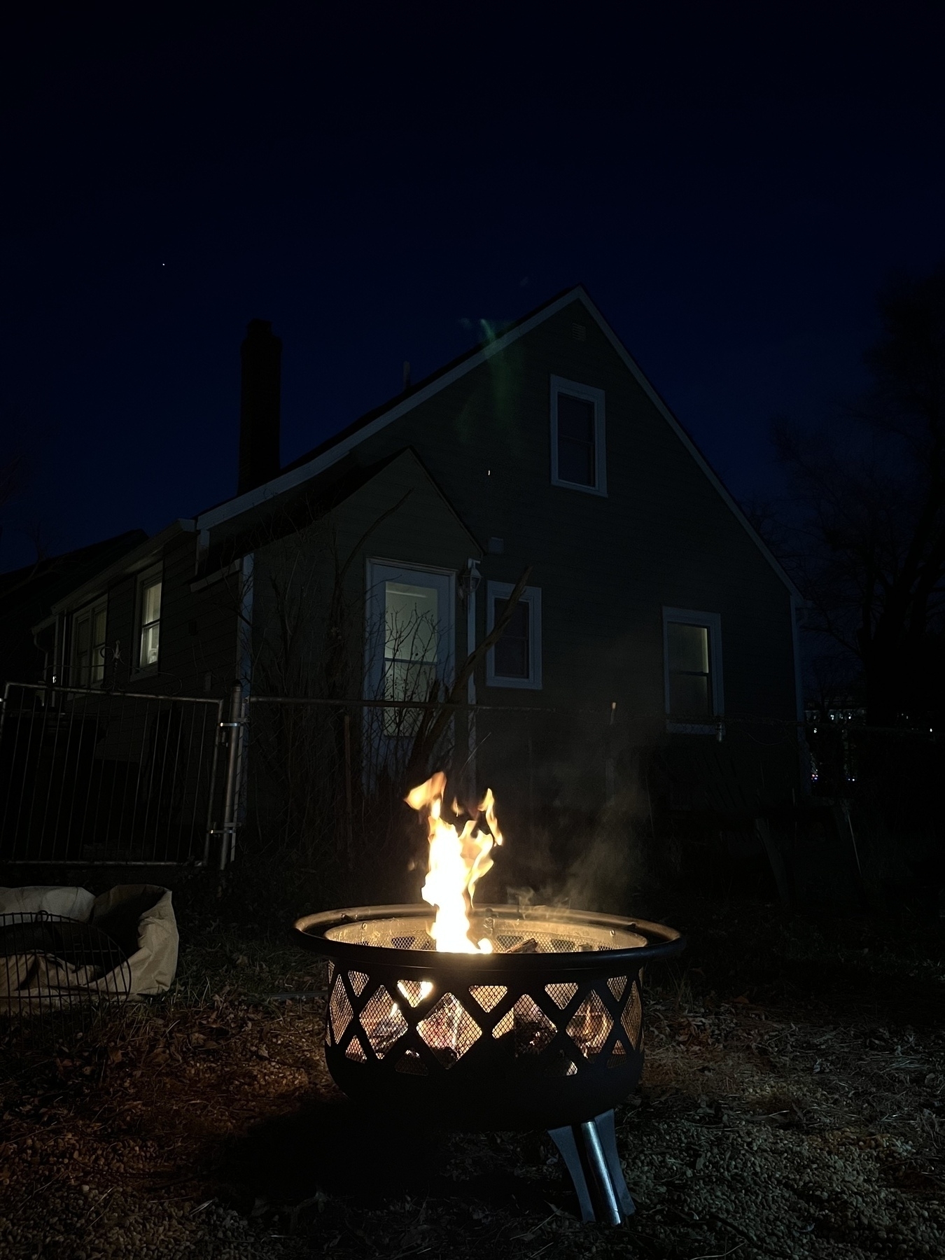 A fire pit with rising flames in a backyard at dusk, with a darkened house in the background and a clear evening sky above.