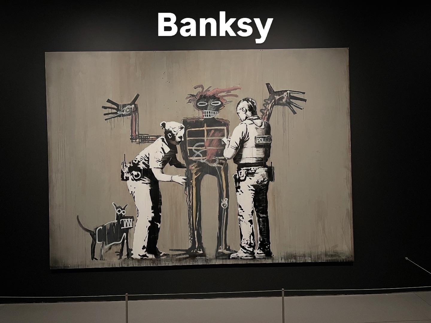 A painting shows two police officers frisking a graffiti-style figure with exaggerated features, while an alert dog stands nearby, set against a plain wall. “Banksy” is written above.