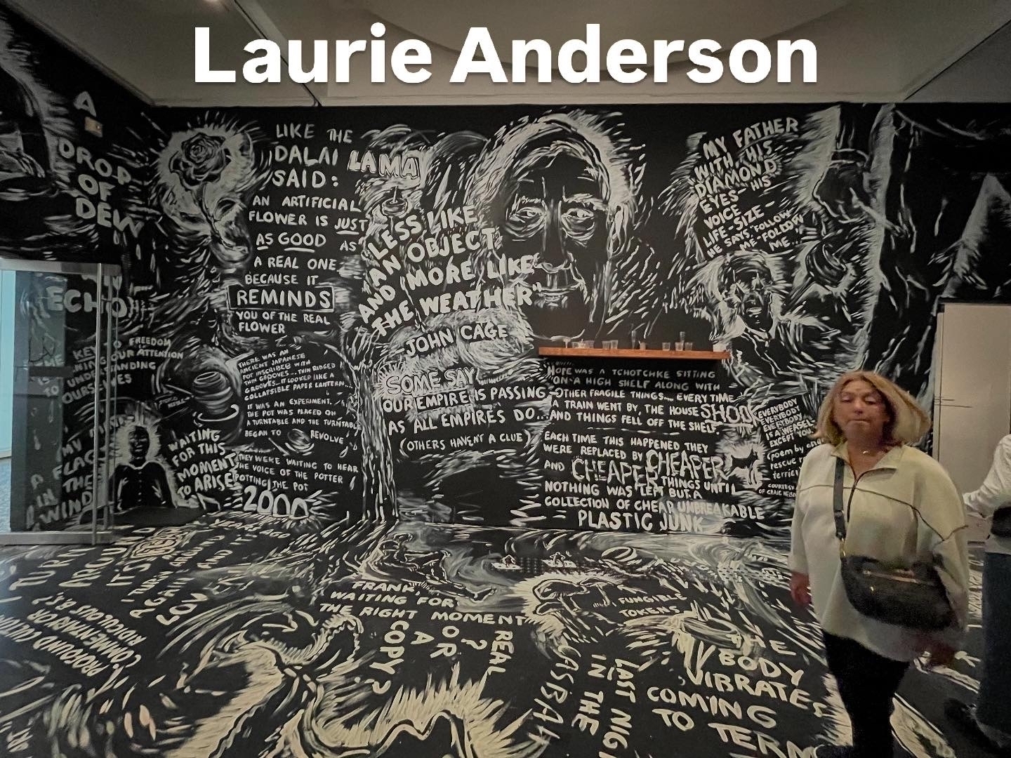 A woman walks through an art installation featuring black and white wall murals with intricate text and drawings. Prominent text includes “Laurie Anderson,” “REMIND,” and “John Cage.