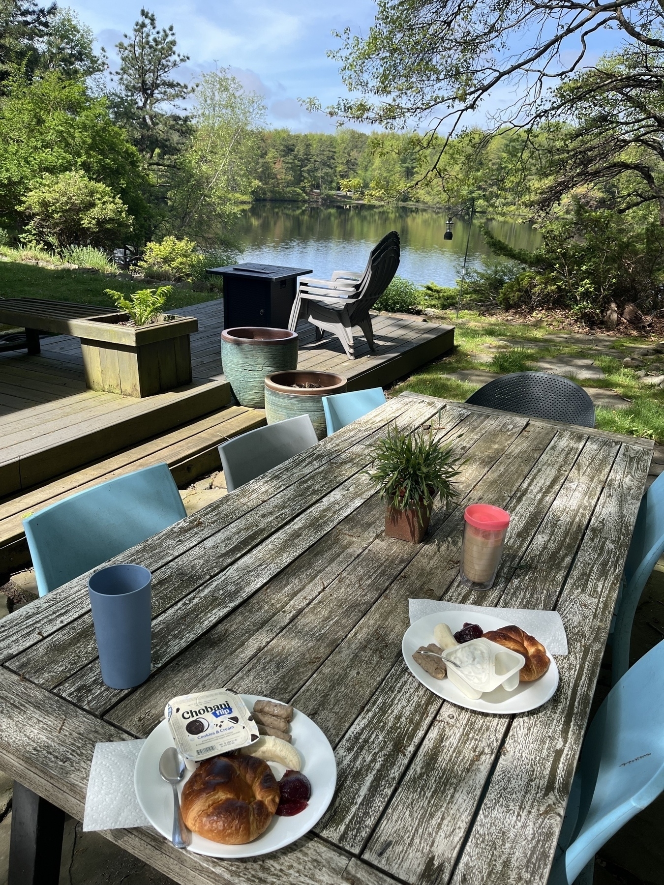 Breakfast plates with croissants, yogurt, bananas, sausages, and jam on a weathered wooden table. Blue chairs surround the table. In the background, a deck with planters, chairs, and a view of a lake and trees under a blue sky.