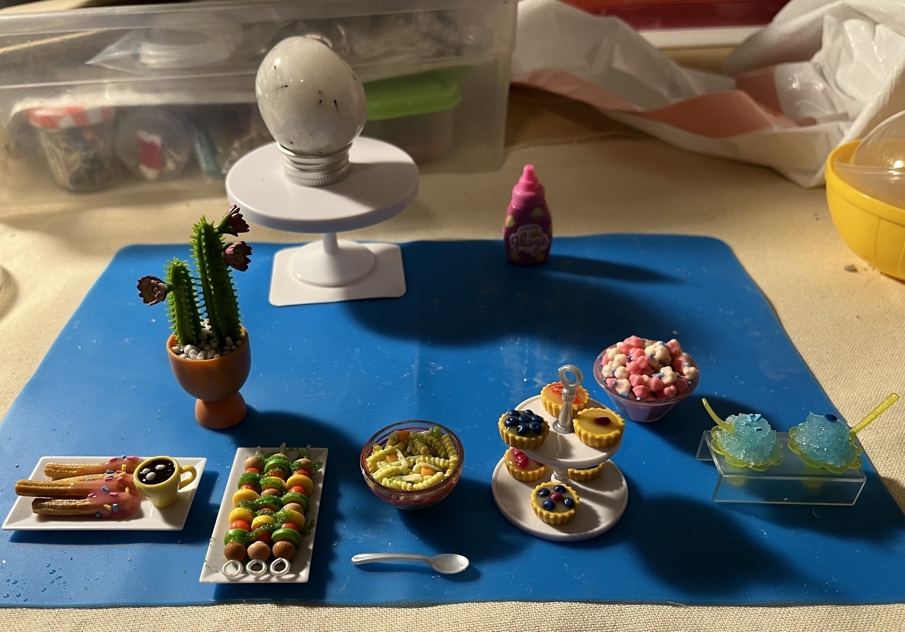 Collection of tiny pastries, churros, popcorn, chicken noodle soup, cactus, snow cones and a crystal sitting on a tiny table in the background. 
