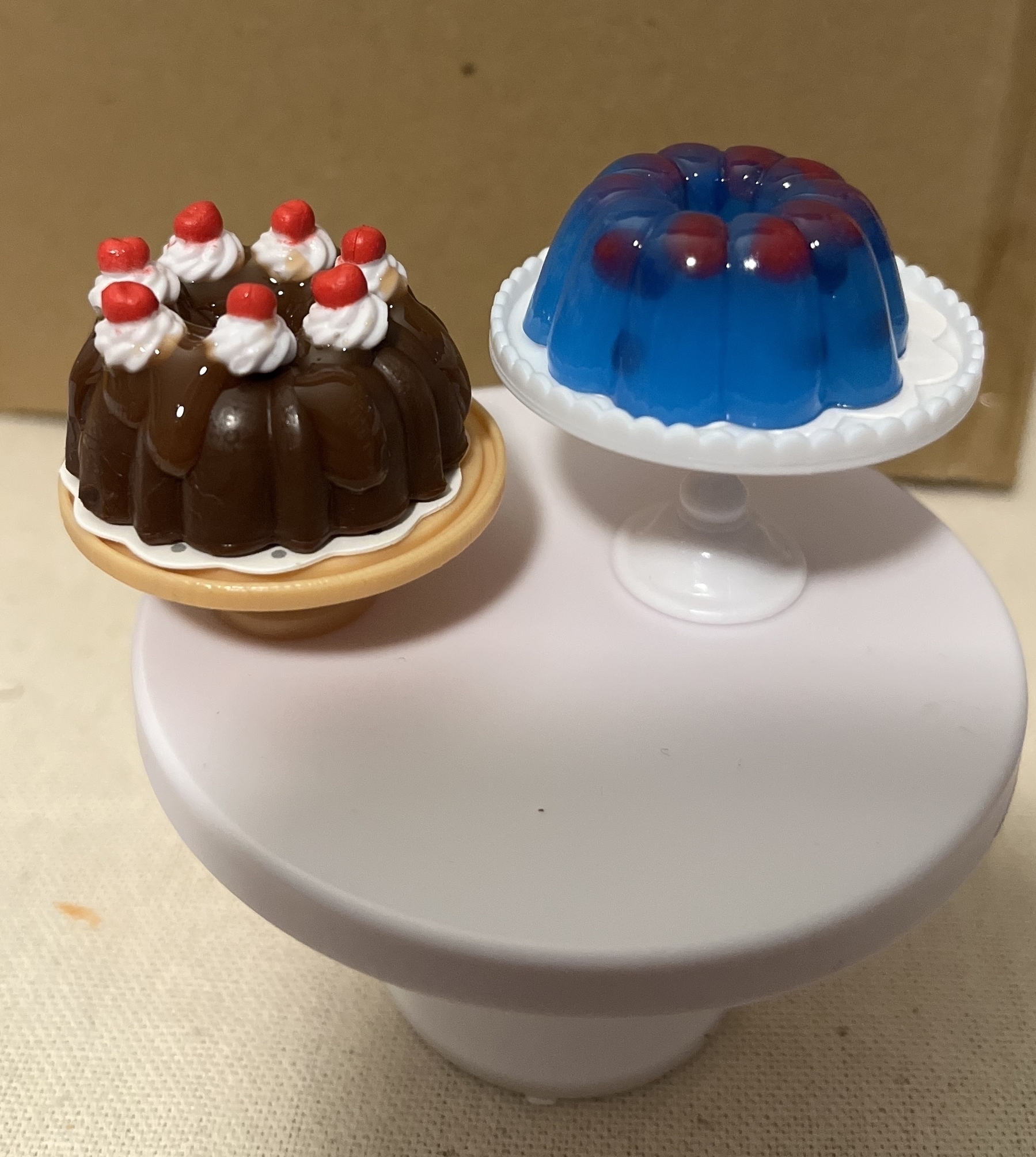 A toy chocolate bundt cake with white icing and red berries on a small plate, next to a toy blue gelatin dessert on a cake stand.