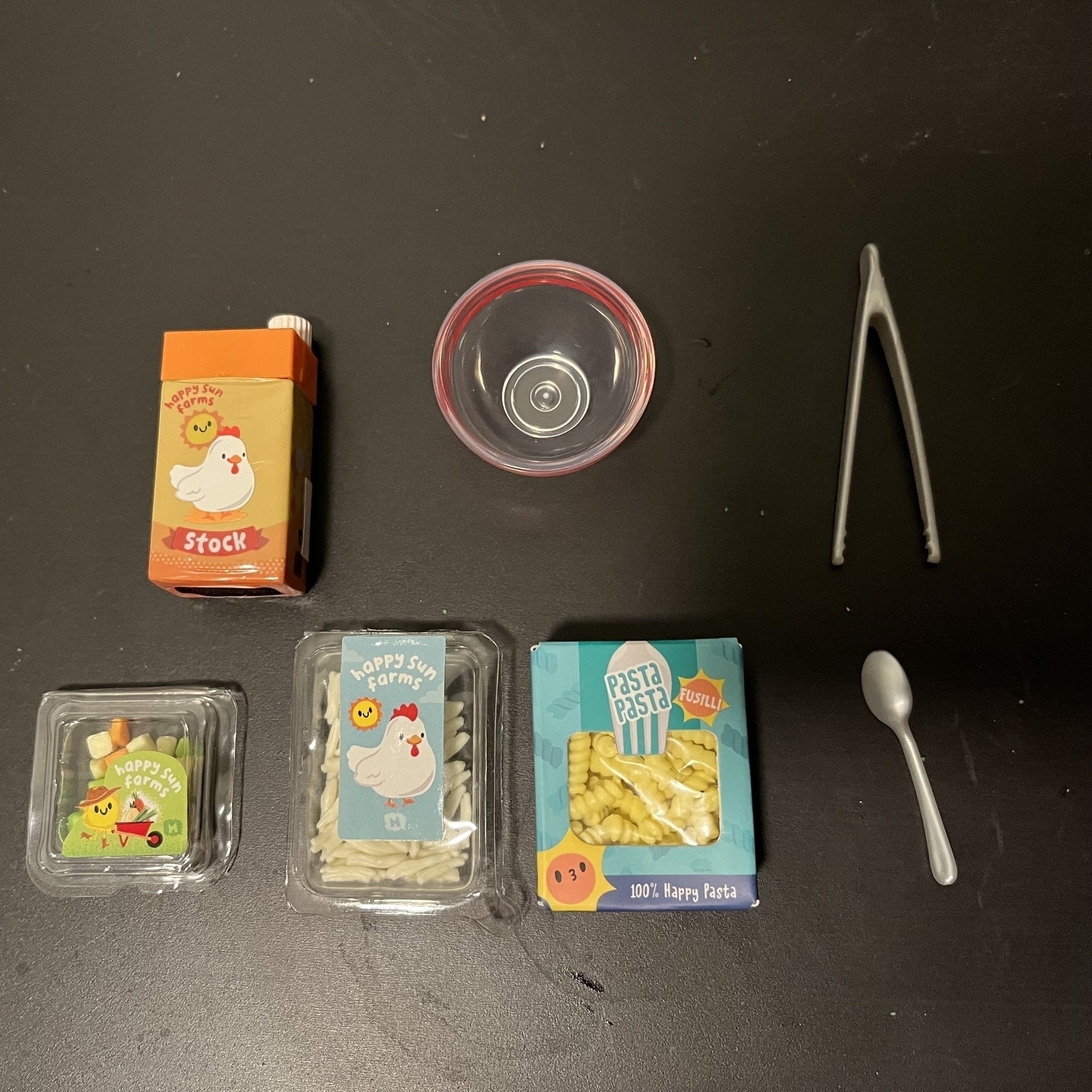 A tiny box of chicken stock holding resin, tiny noodles, chicken strips, vegetable cubes, an empty bowl and tweezers and a spoon. 