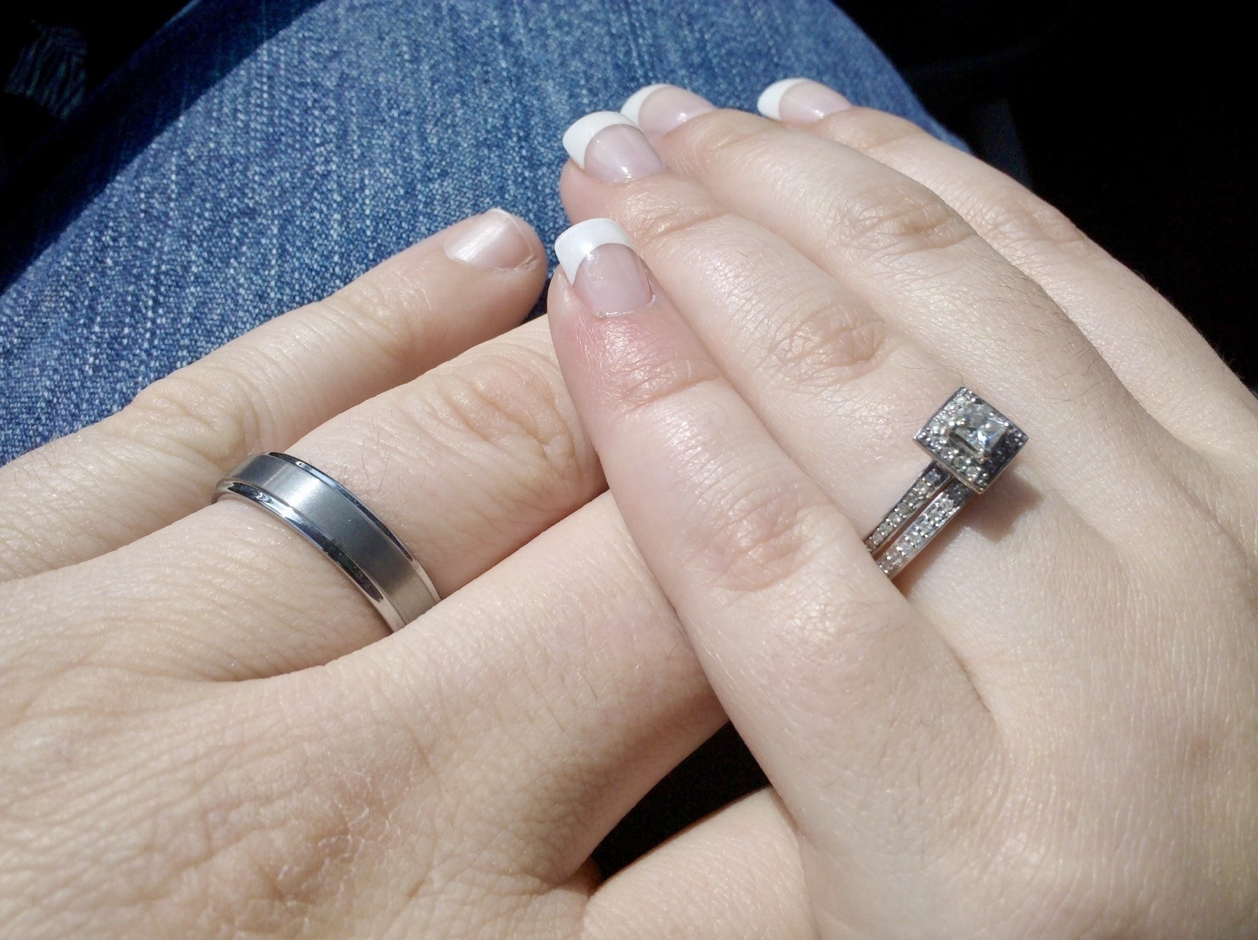 Two hands with wedding rings rest on a denim-clad leg. The left hand has a simple metal band, while the right hand features a square-cut diamond ring with a matching band. The right hand has manicured nails with French tips.