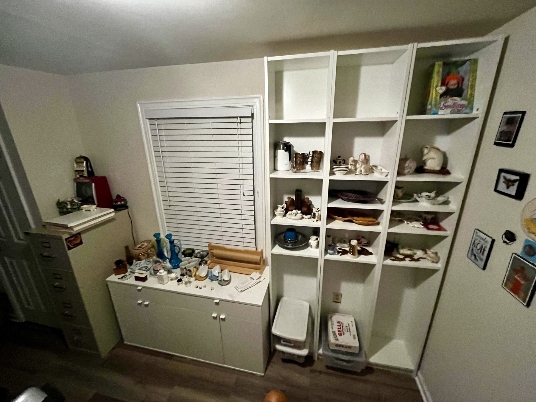 A cluttered room corner with a filled white shelving unit, objects on a windowsill, artwork on walls, and a filing cabinet to the left.