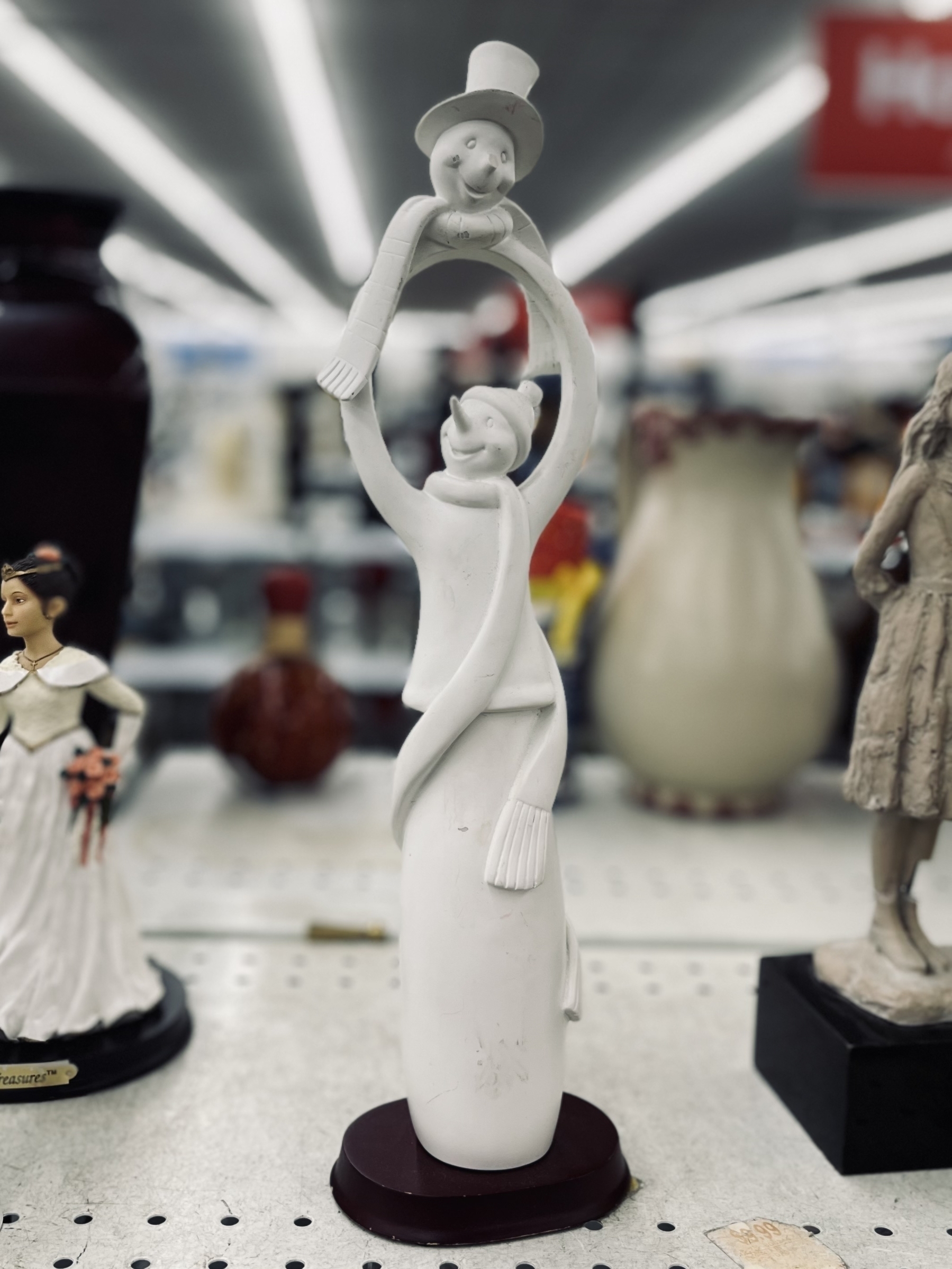 A white snowman figurine depicts two smiling people in hats holding hands and forming an arch, displayed on a store shelf. 