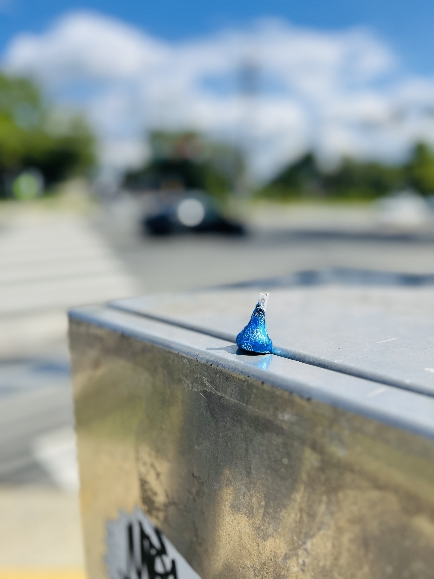 A single blue Hershey’s Kiss candy wrapper rests on a metallic ledge against a blurred urban backdrop with foliage and passing cars.