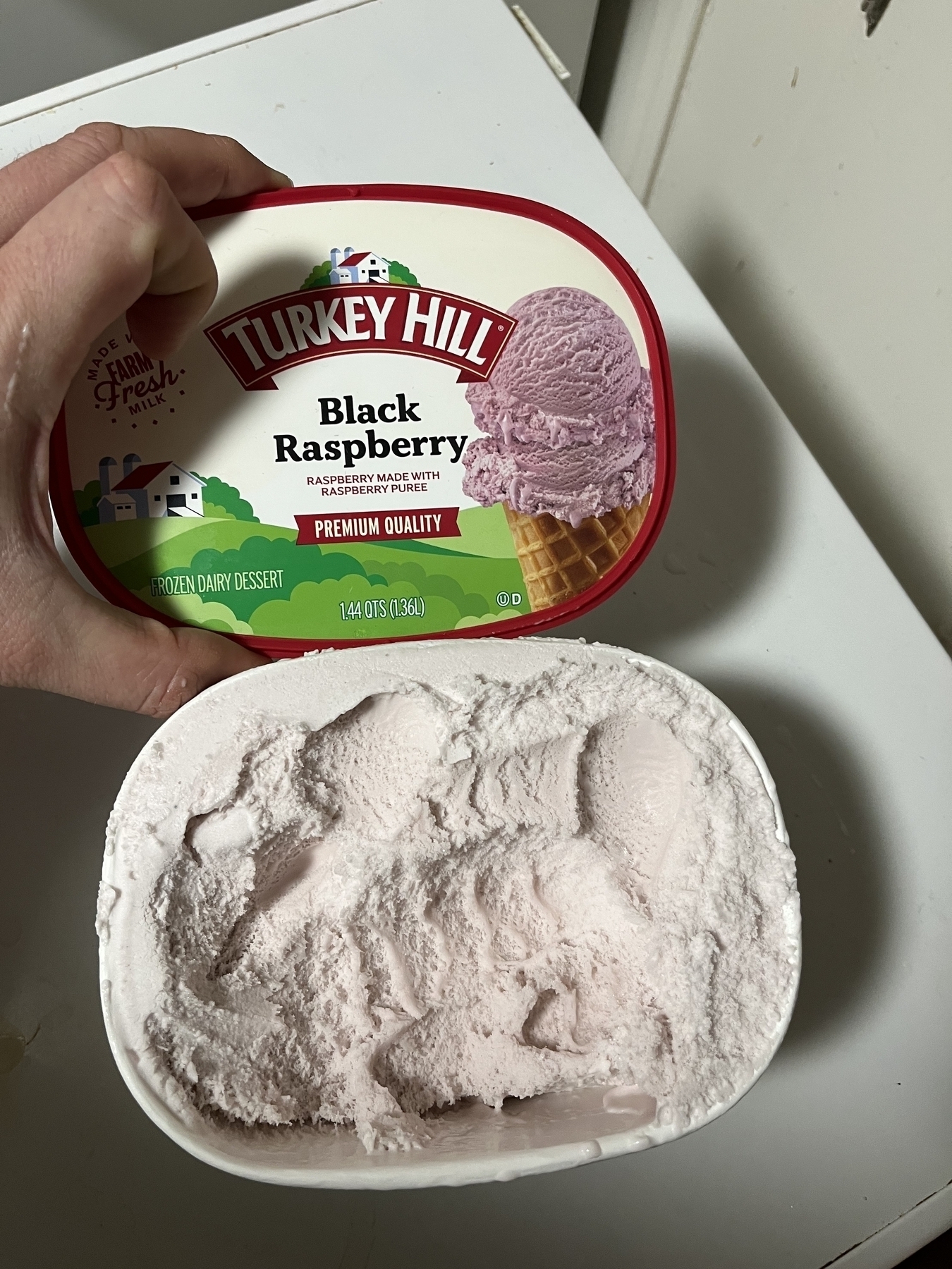 A hand holds an open carton of Turkey Hill Black Raspberry frozen dairy dessert, partially scooped, atop a white surface.