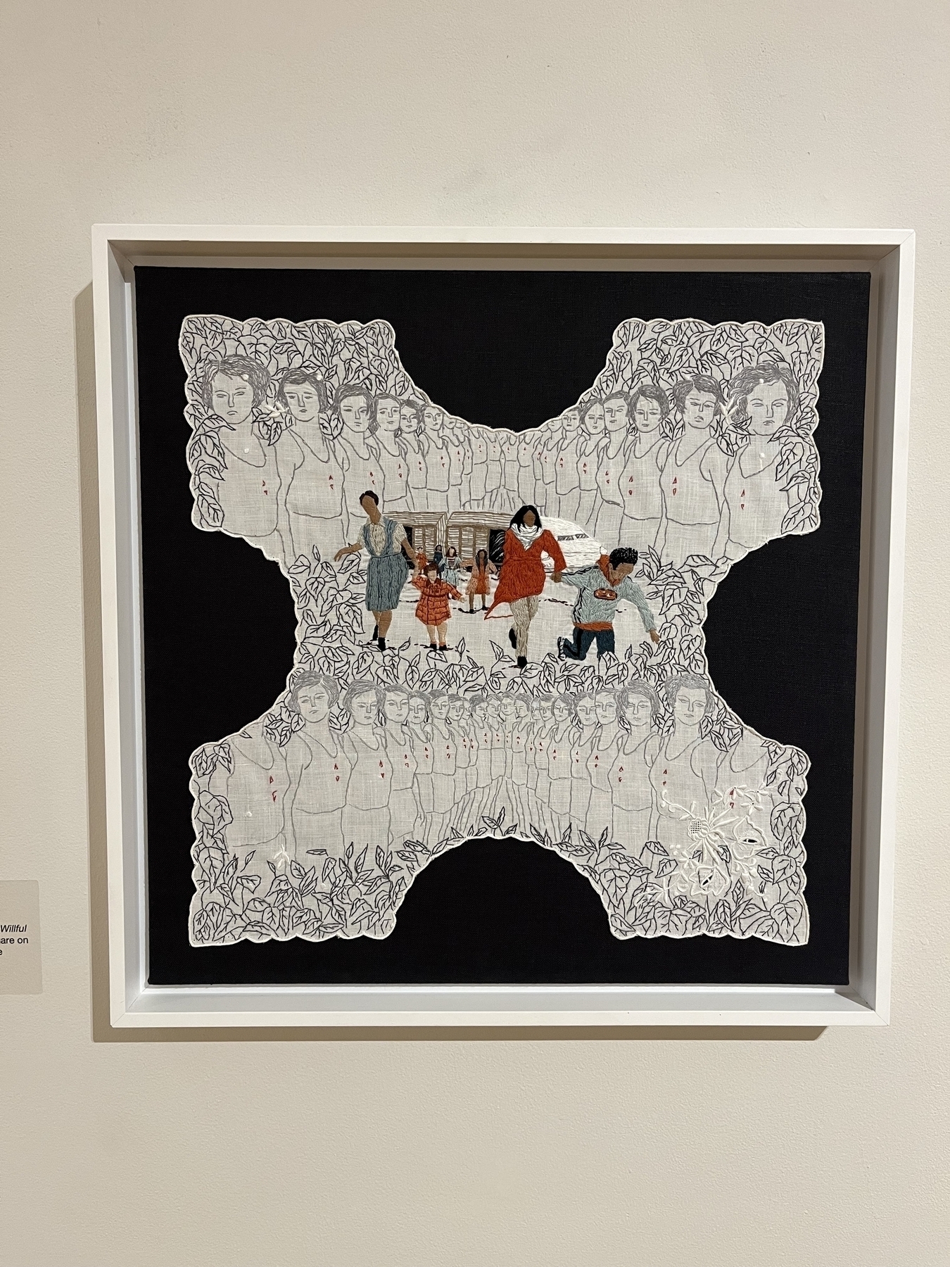 A framed textile artwork depicting illustrated people with a central colorful group, displayed on a gallery wall.