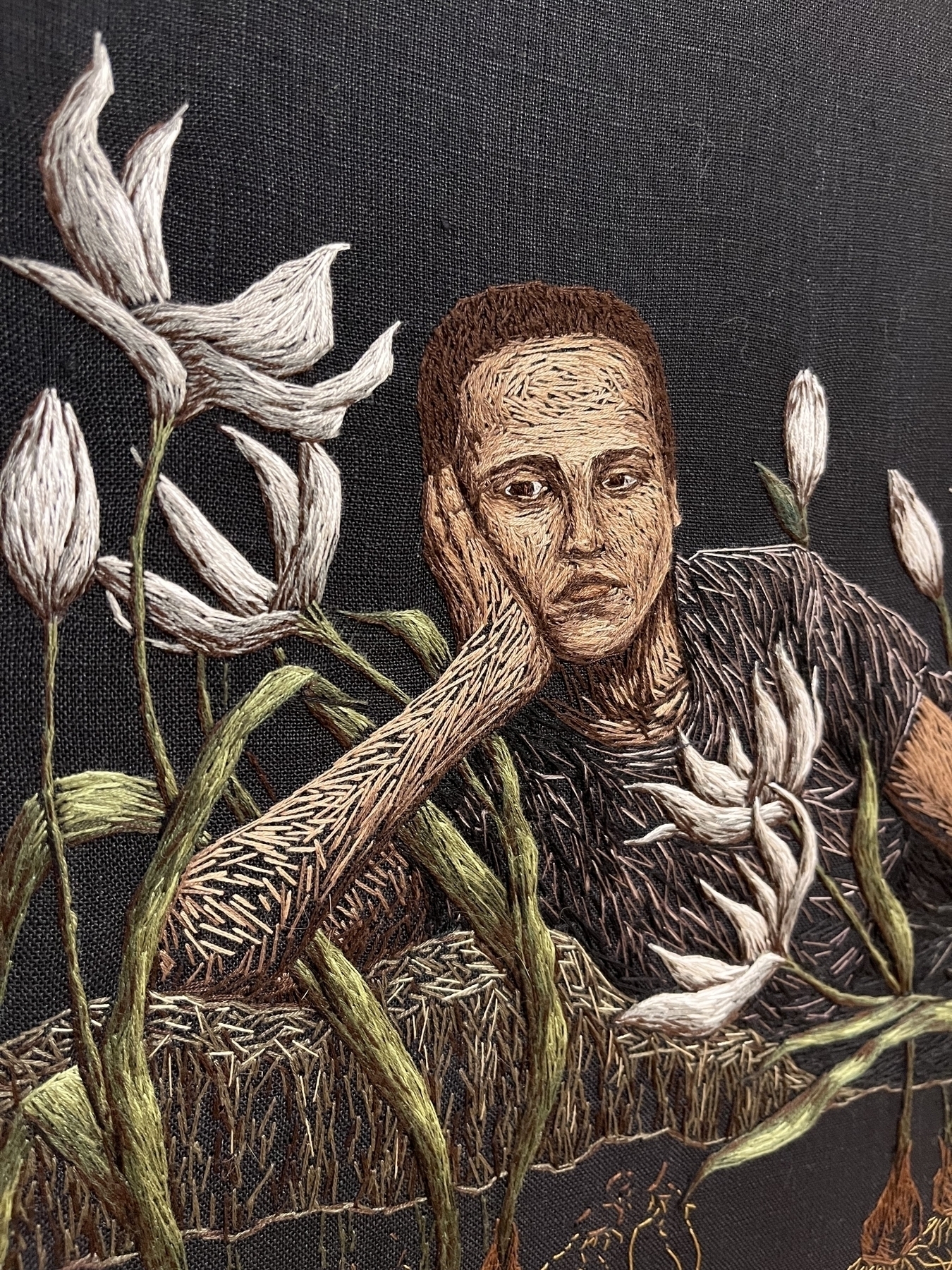 A person rests their head on their hand, embroidered with thread among white lilies on a black background, creating a textured, contemplative scene.