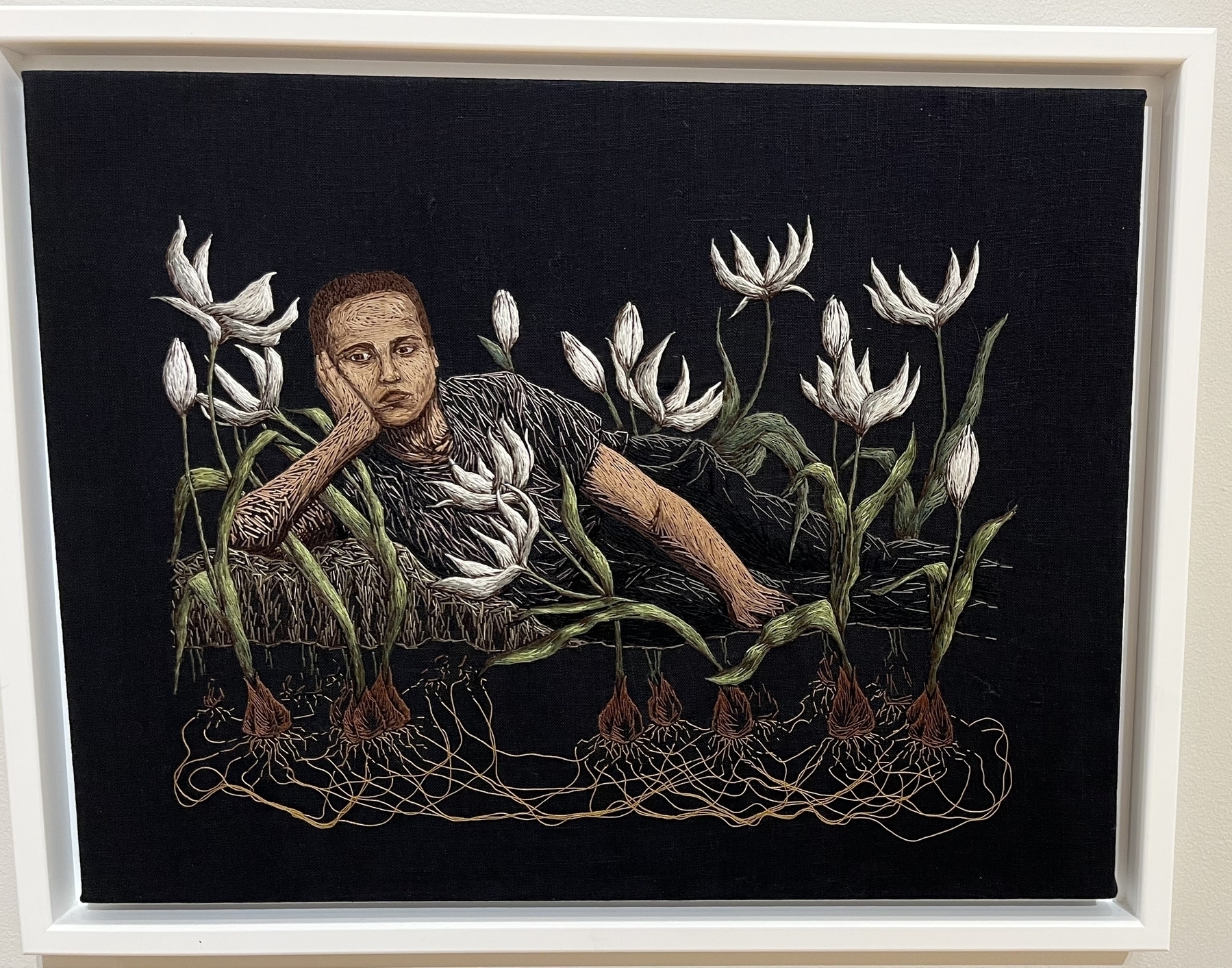 Embroidered figure rests amongst white flowers on a dark background, conveying introspection or melancholy, framed for display.