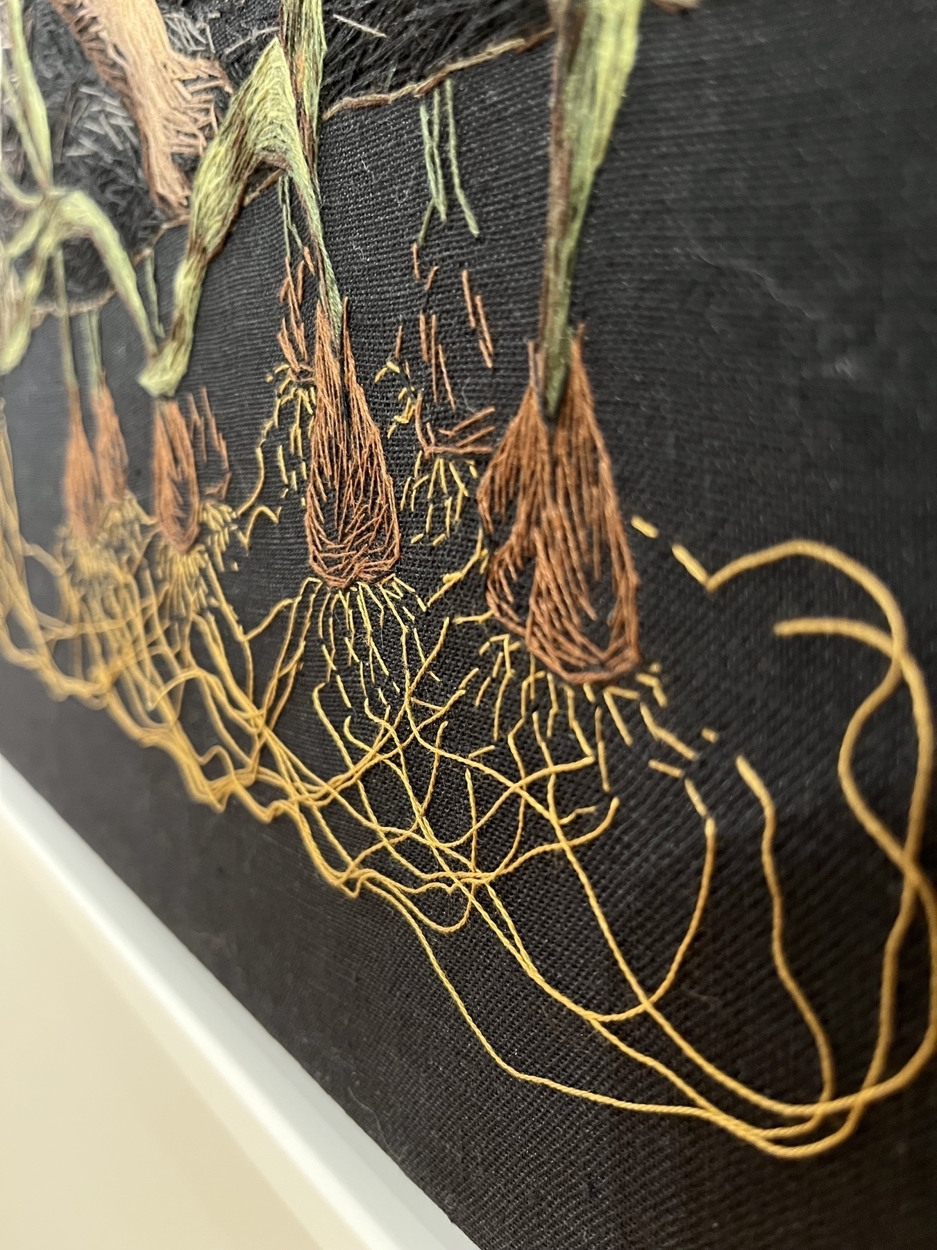 Embroidered artwork depicting textured plant roots in various colors against a dark background, giving a detailed close-up perspective.