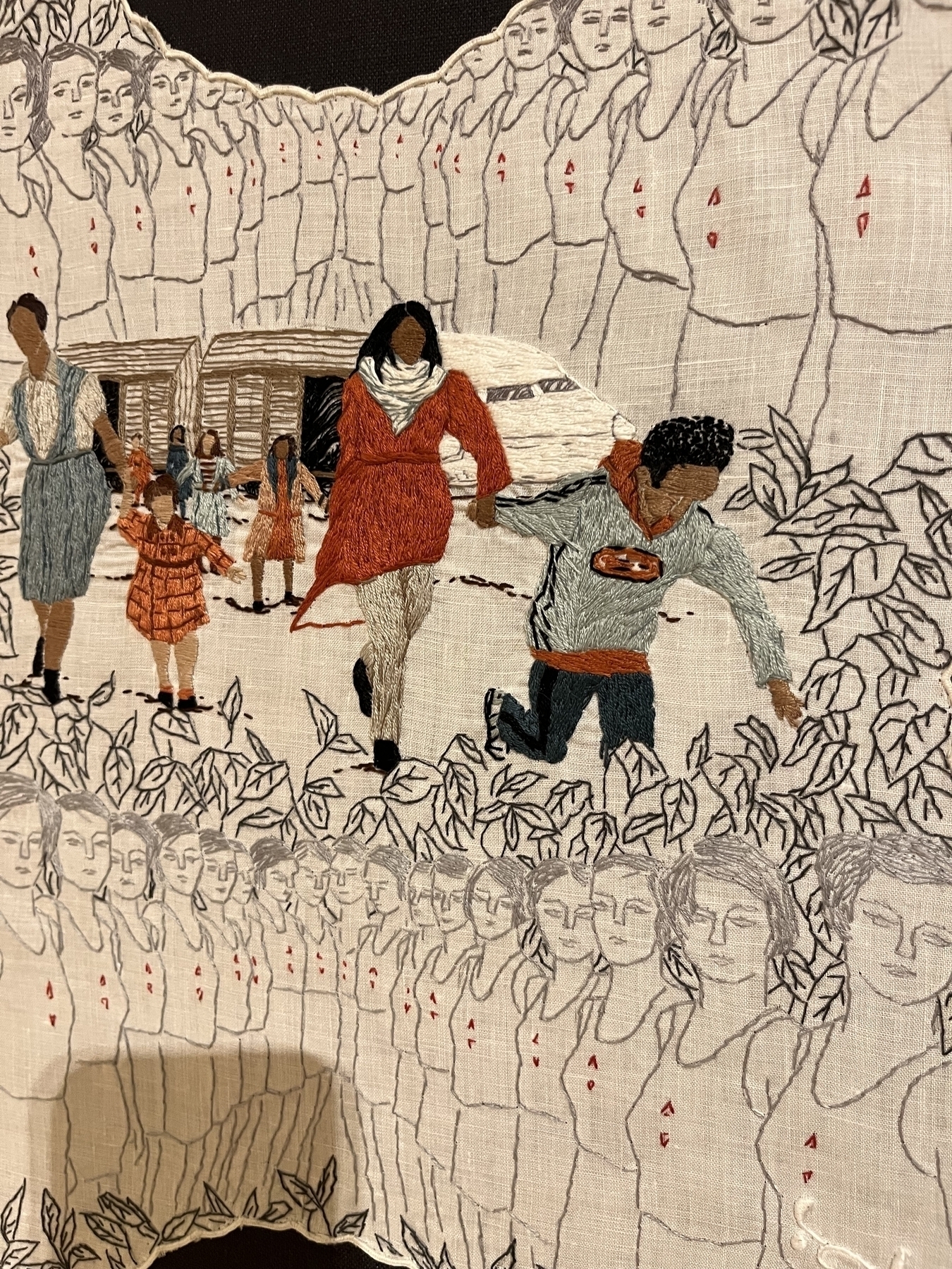 Embroidered figures engage in varied activities against a backdrop of more figures and foliage, suggesting a community scene on fabric.