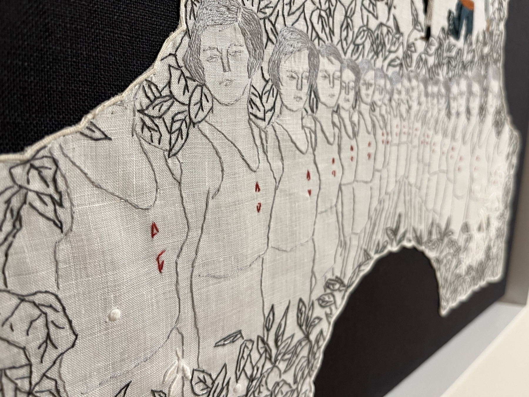 Embroidered figures on fabric, with repeating pattern of women against a floral backdrop, displayed partially over a black background, suggesting depth and texture.