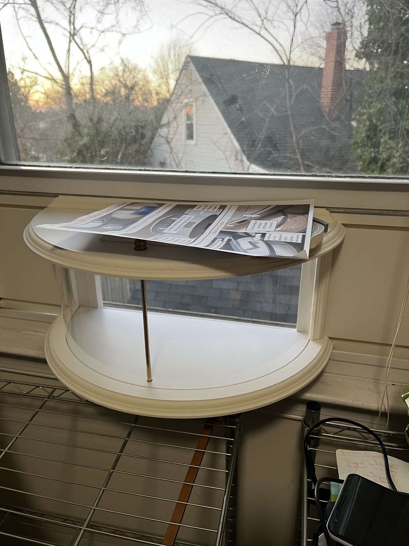 A two-tiered, oval window bird feeder holds assorted papers, set against a sunset view through a screened window, with a silhouette of a house outside.