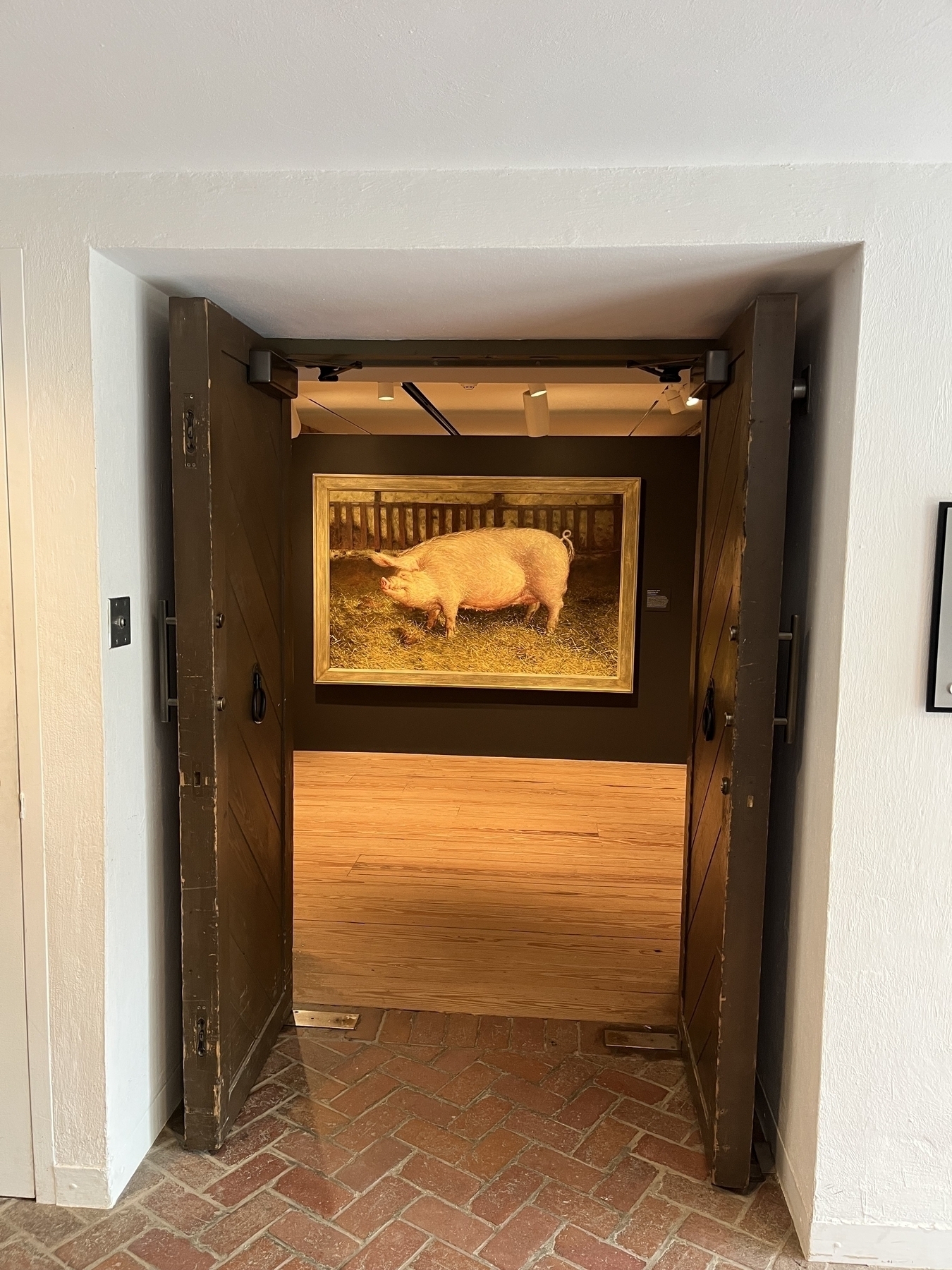 Painting of a pig hangs on a wall, illuminated by spotlight. Viewed through a doorway with brick floor tiles and wooden doors.