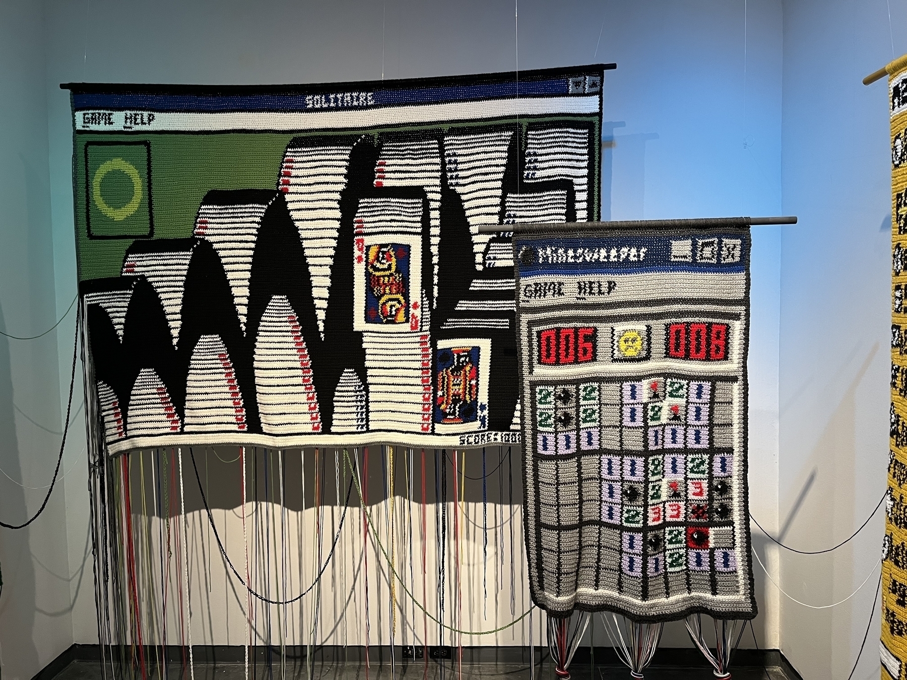 Two woven tapestries depict pixelated computer game screens of minesweeper and solitaire. The larger one shows a Solitaire game with stacked cards; the smaller one resembles Minesweeper with numbered blocks.