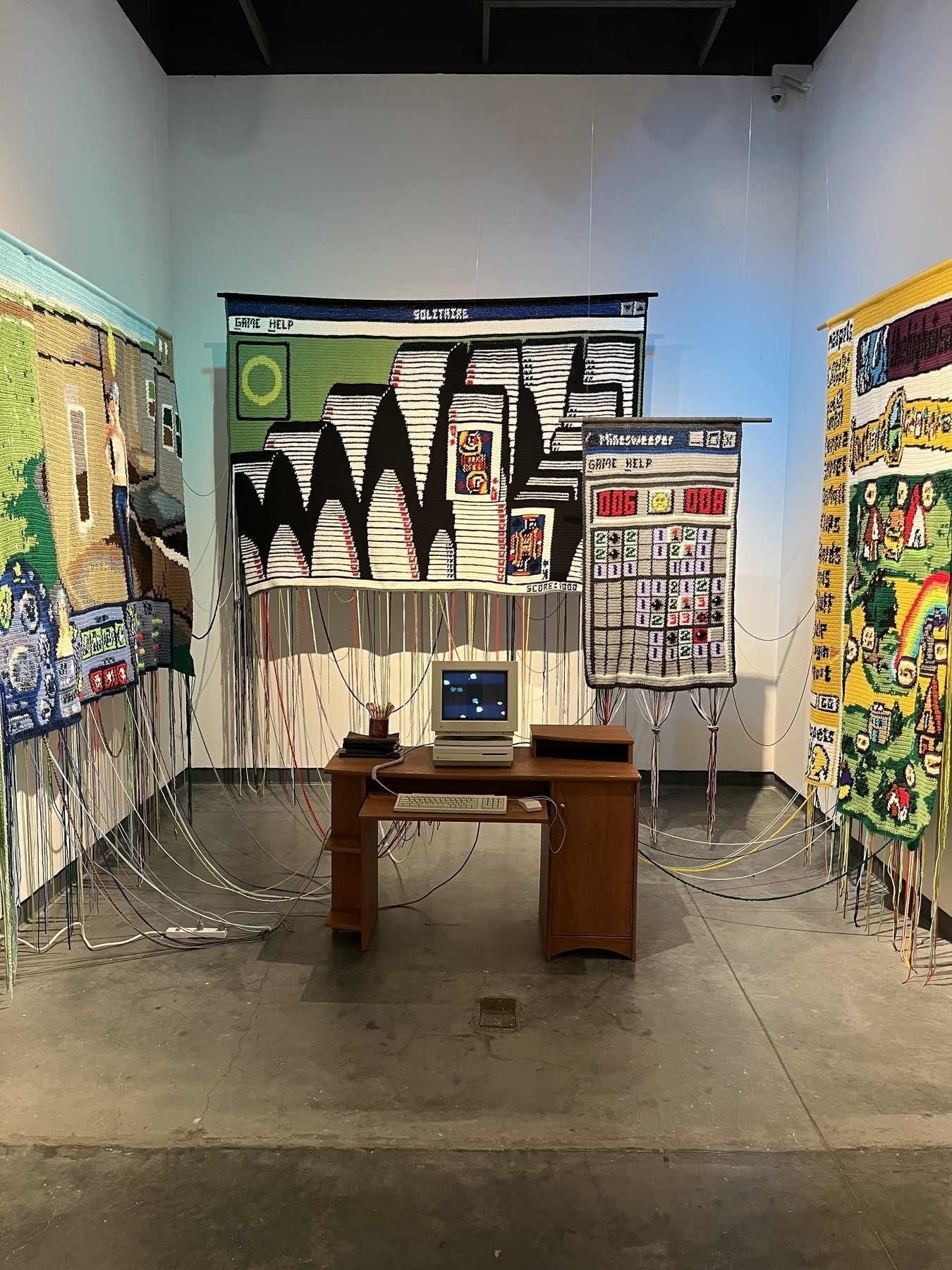 A vintage Macintosh computer sits on a wooden desk displaying flying toasters. Woven tapestries depict minesweeper, the Sims 2, neopets and solitaire on surrounding walls, with colorful threads creating a networked effect throughout the gallery space.